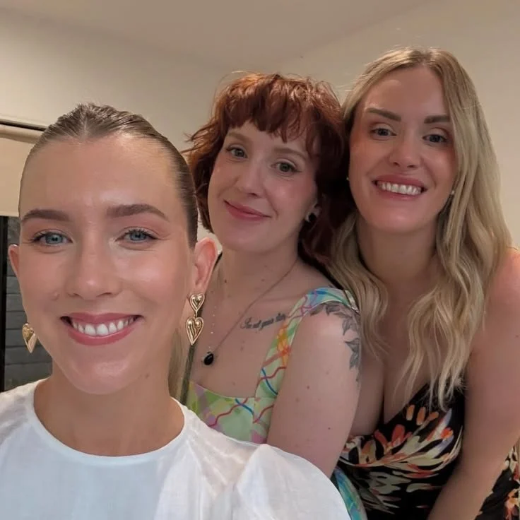 Three women smiling, all with soft glam makeup, taking a selfie together indoors.
