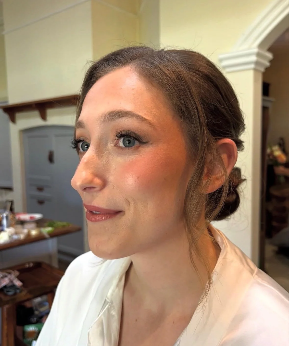 Close-up of a bride with styled hair, natural glam makeup, and wearing a white garment, in a softly lit indoor setting.