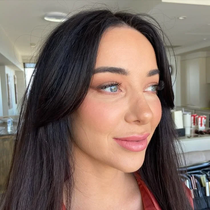 Close-up portrait of a woman with long dark hair, blue eyes, and natural-glam makeup, smiling softly in a well-lit indoor setting.