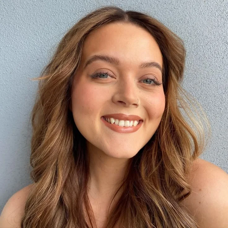 Close-up of a confident young woman with natural glam makeup and wavy brown, hair smiling in front of a light blue wall.