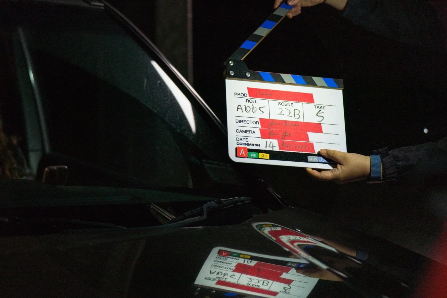 A film director's clapboard being held by a person for a scene in a dark setting, with a black car in the background.