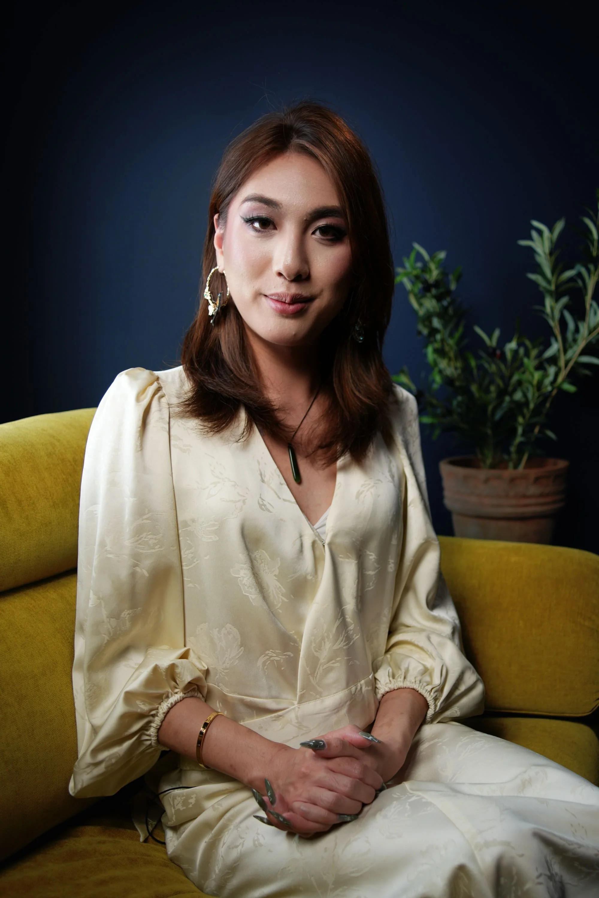 A woman with shoulder-length brown hair, wearing earrings and a cream-colored satin blouse, sitting on a yellow couch with a potted plant in the background.