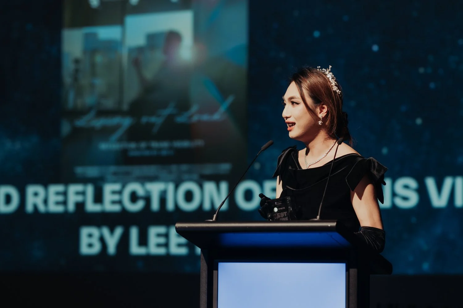 A woman in a black dress giving a speech at a podium during a conference or presentation, with a large screen behind her displaying a presentation slide.