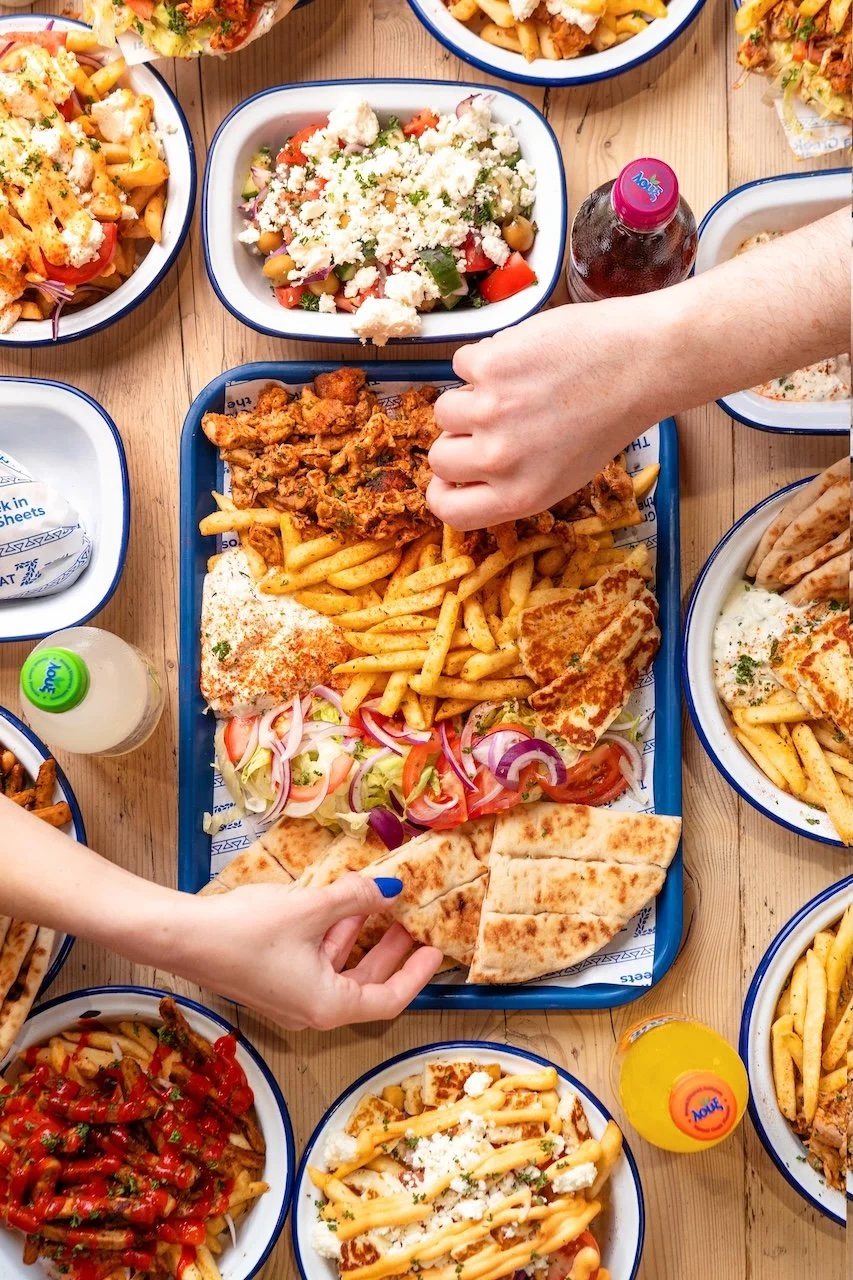 A variety of Mexican dishes on a wooden table, including tacos, French fries, salads, and drinks, with hands reaching for food.