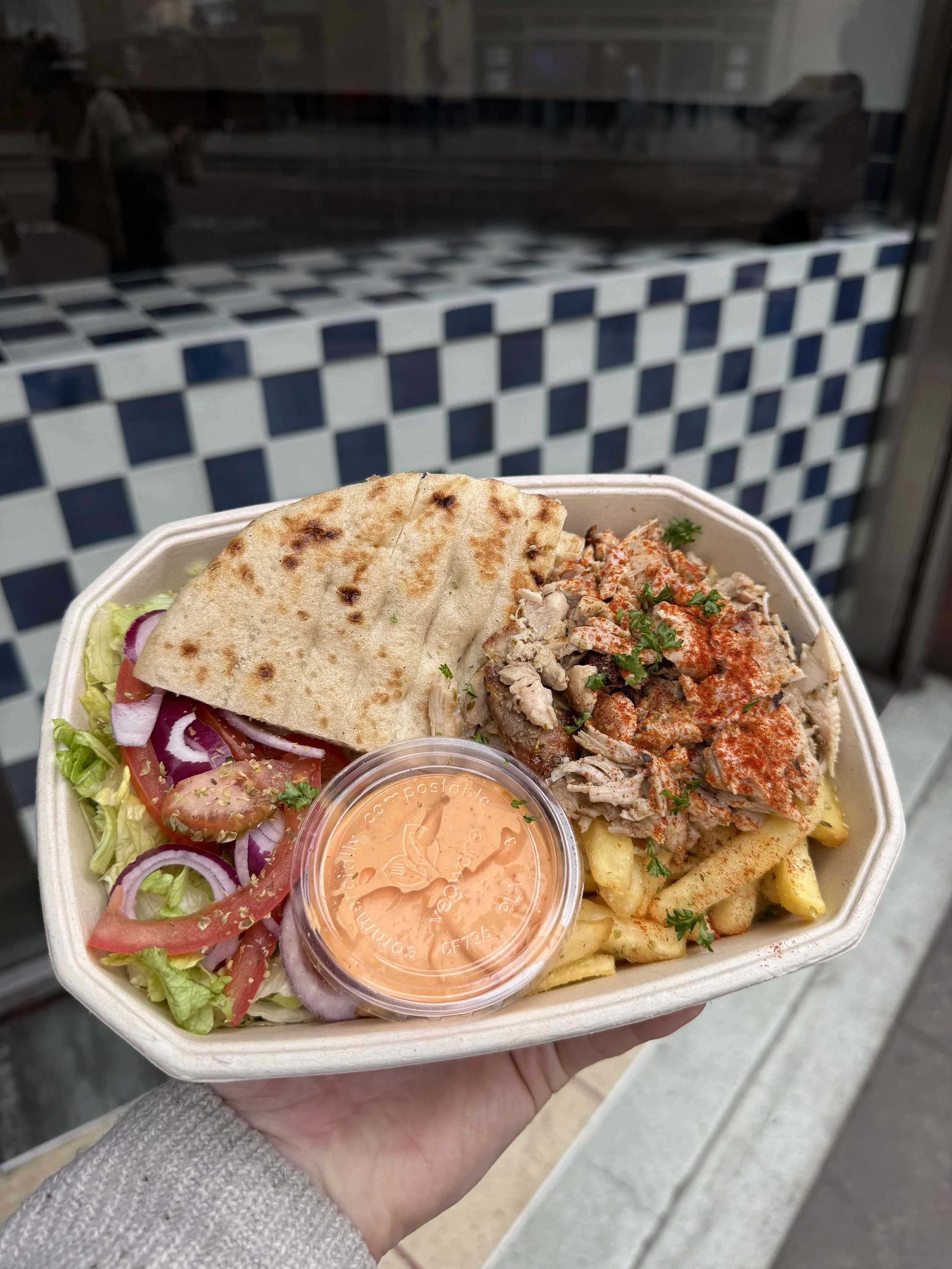 A takeout container filled with salad, pita bread, grilled chicken, French fries, and a small cup of pink sauce, held by a person outside near a tiled wall.
