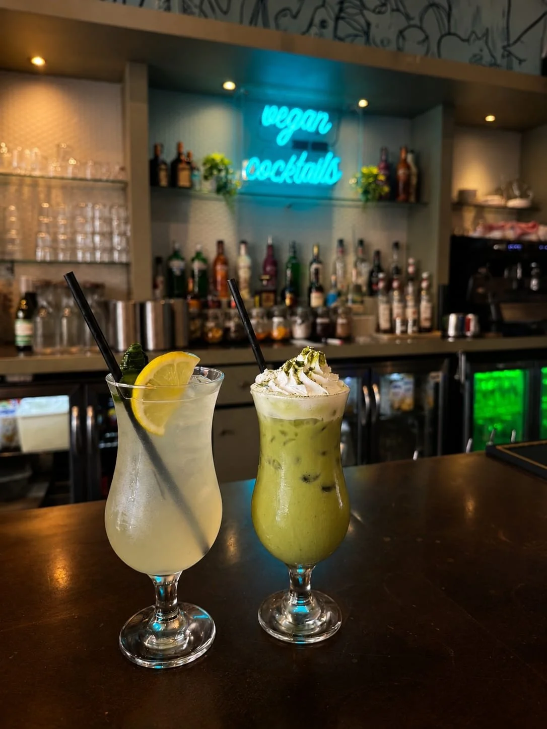 Two cocktails on a dark bar counter in front of a shelves stocked with bottles of alcohol. One is a pale yellow drink with a lemon wedge and mint leaf garnishes, the other is a green drink topped with whipped cream and a drizzle of green syrup. A neon sign in the background reads 'vegan cocktails'.