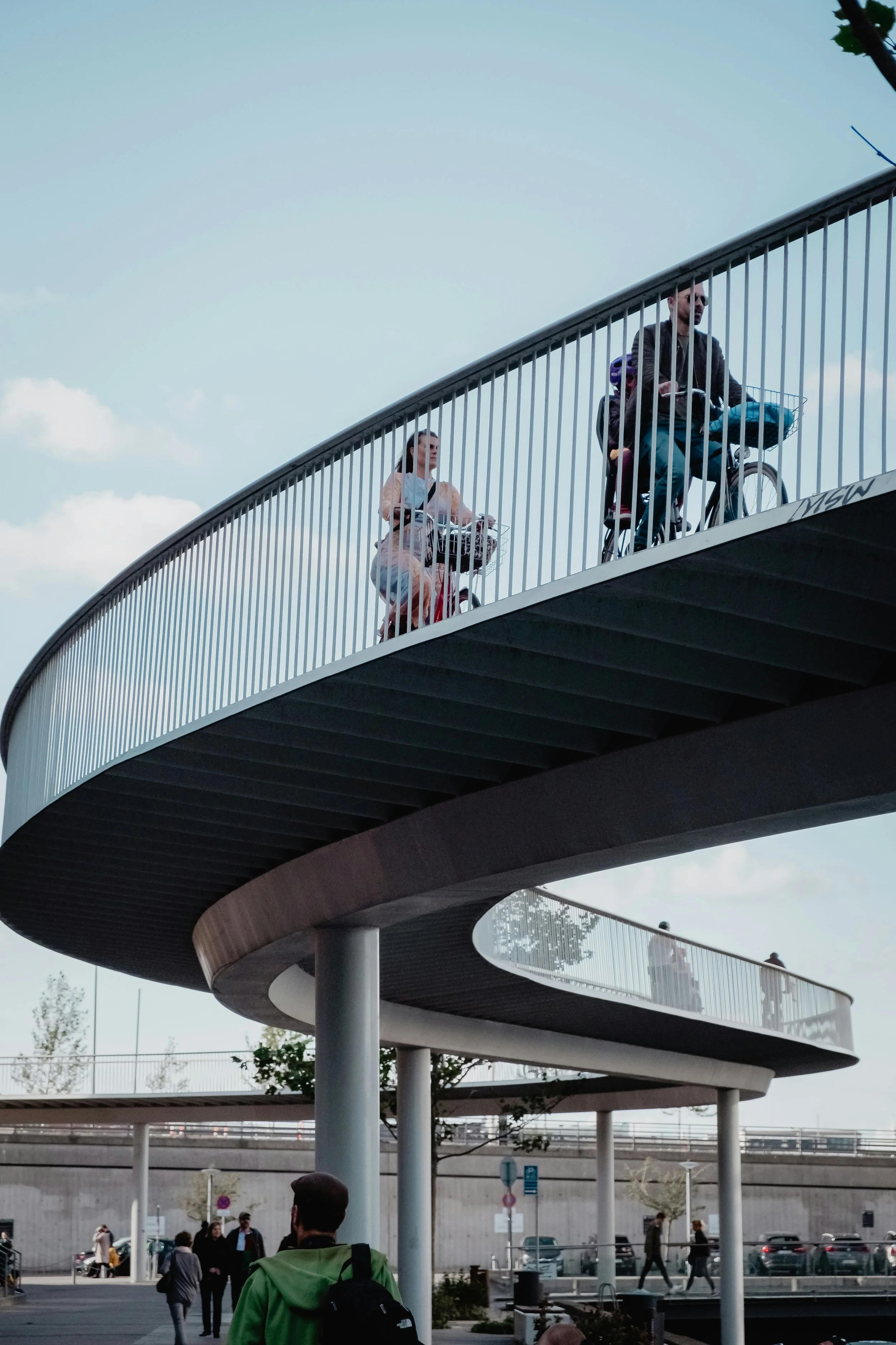 People cycling on a curved, modern pedestrian bridge with vertical railings, blue sky, and trees in the background.