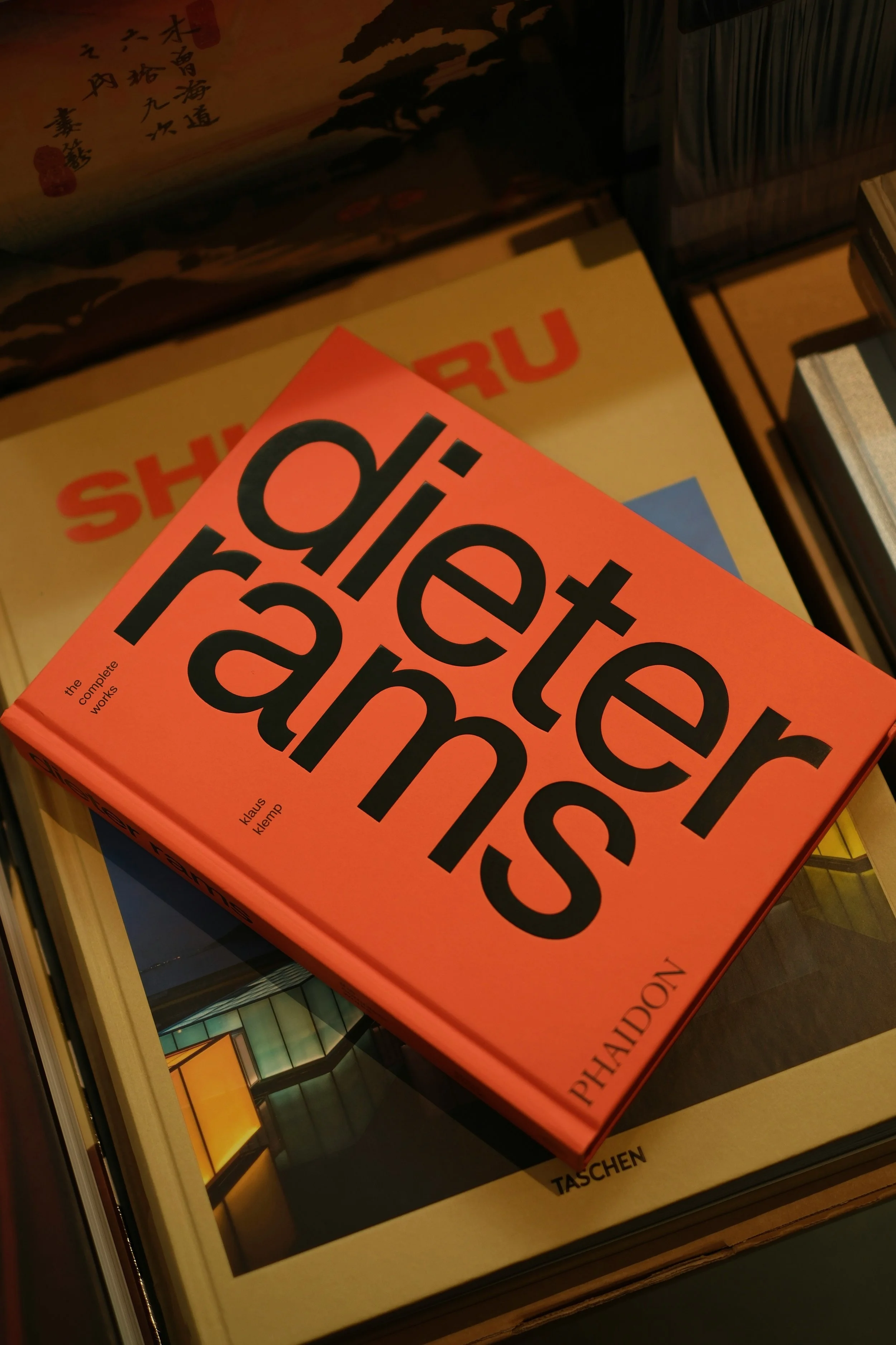 A stack of books, with one orange book of Dieter Rams work in the foreground. Other books are partially visible behind it.