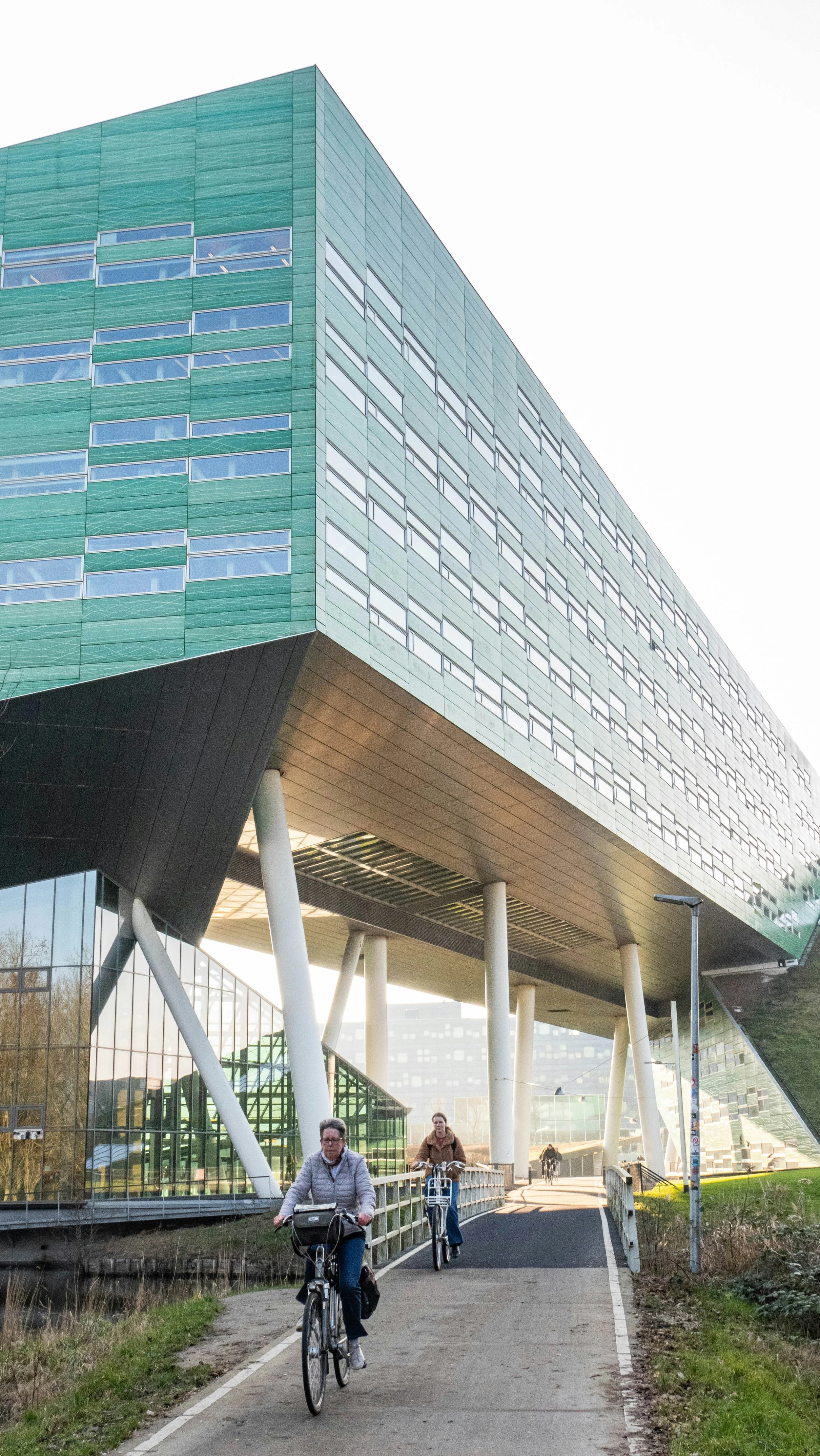 People riding bikes on a designated bike path in front of a modern, multi-story building with glass windows and green exterior panels, supported by large white columns.