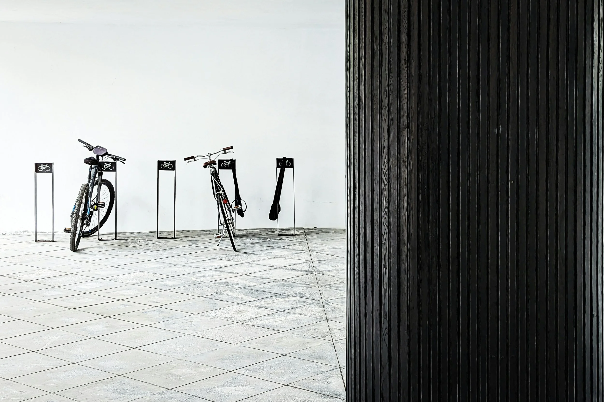 Three bicycles parked at a bike rack against a white wall, with a black vertical wooden wall on the right side.