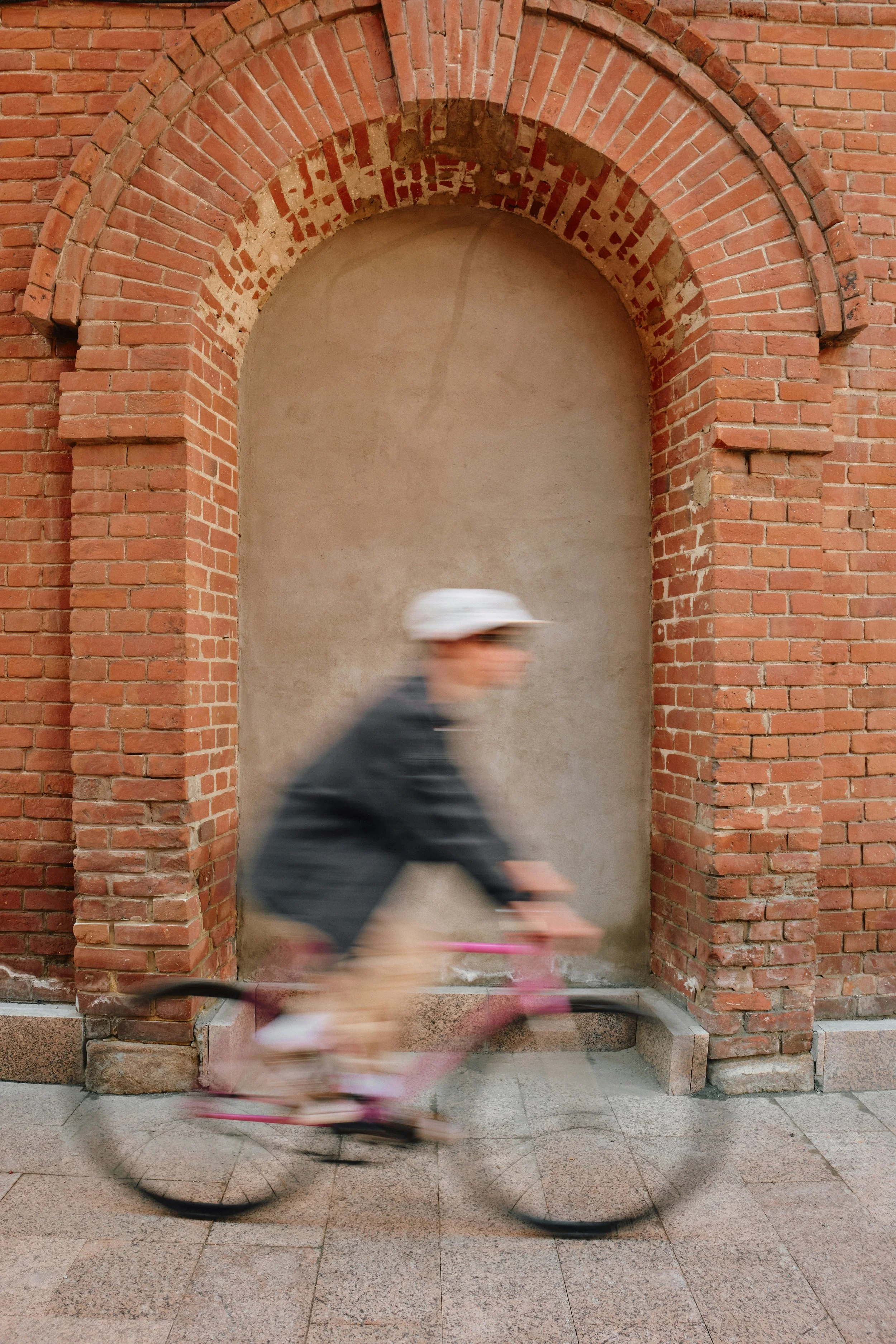 A person riding a bicycle past a brick archway on a sidewalk, with motion blur indicating movement.