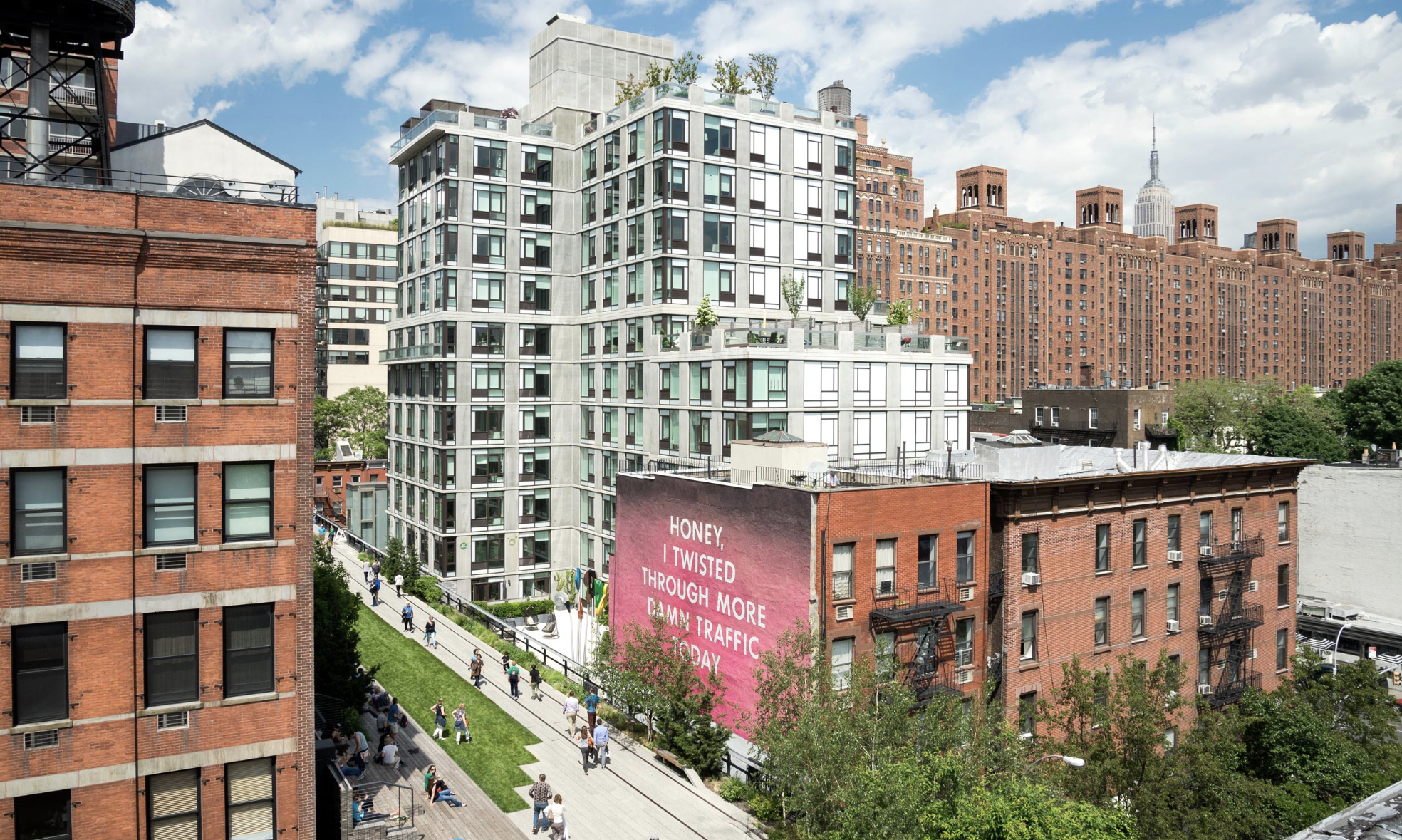Cityscape featuring a modern white apartment building with balconies and greenery on top, surrounded by red brick buildings and a busy pedestrian street below.