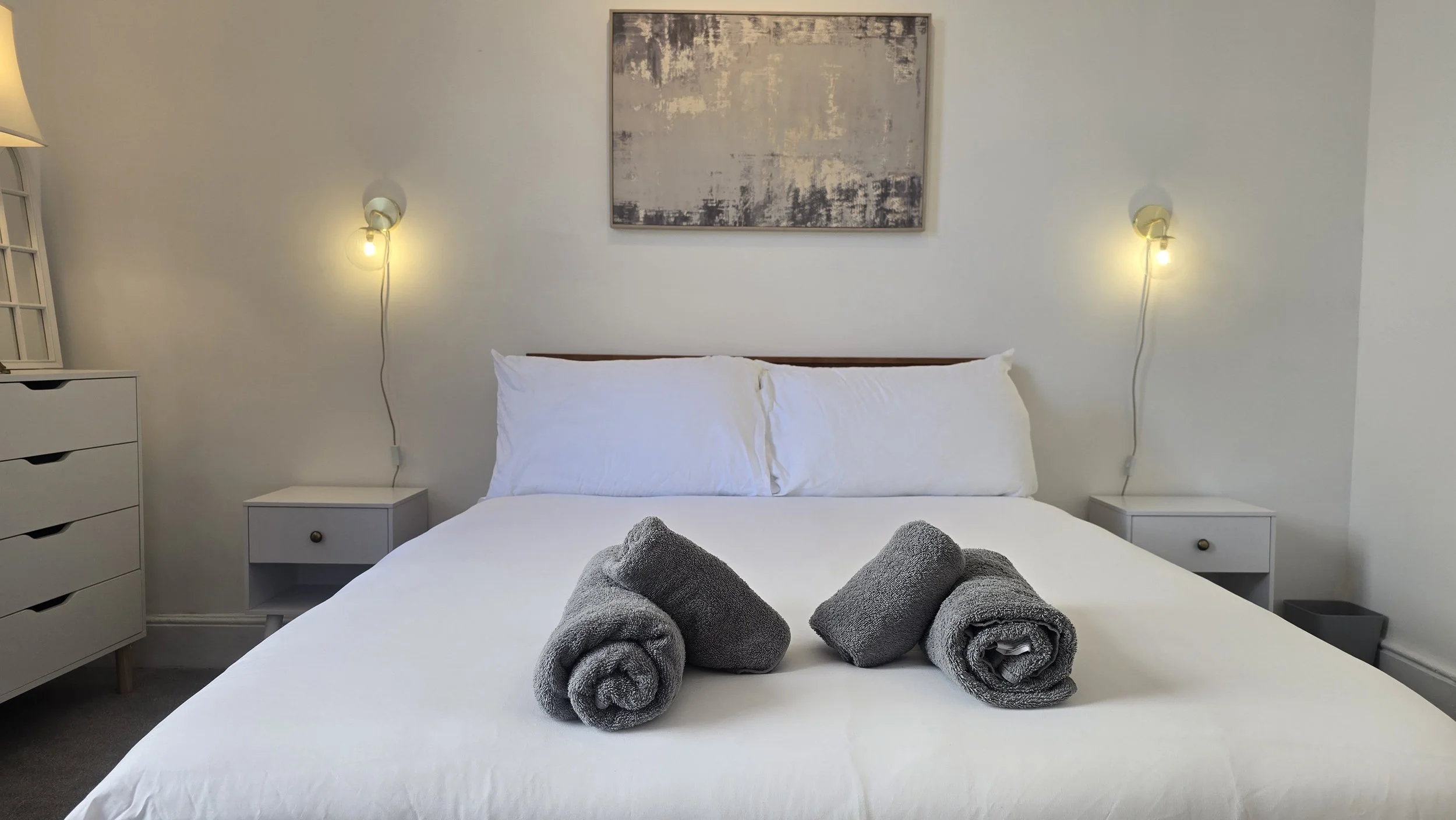 A neatly made bed with white bedding and pillows, with three gray towels rolled up and placed on top. There are two small white nightstands on either side of the bed, each with a drawer, and a modern abstract painting hanging on the wall above the head of the bed. The wall features two wall-mounted lamps with exposed bulbs on either side of the painting, providing warm lighting to the room.