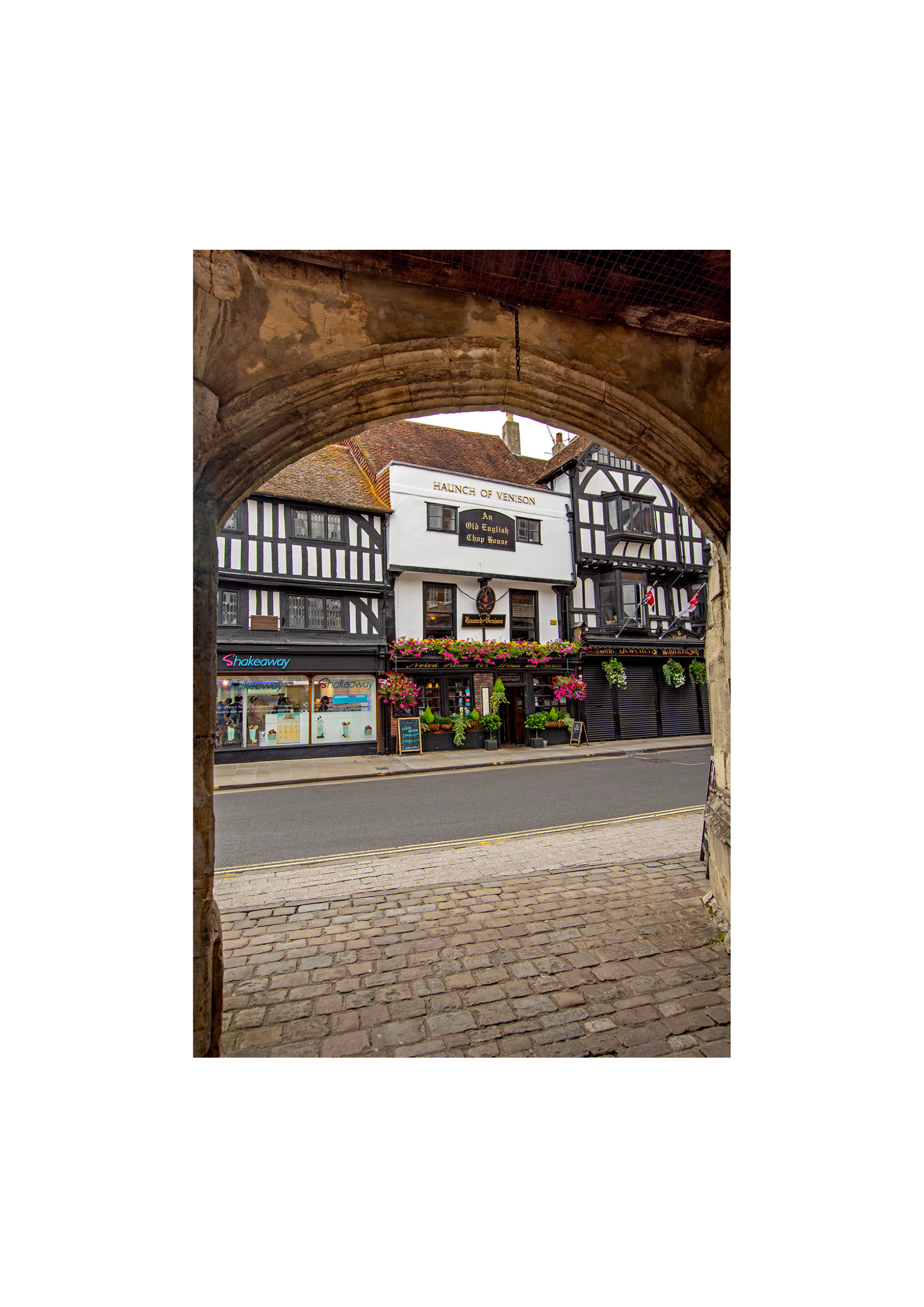 View of a street through a stone archway, featuring a historic black-and-white timber-framed building with a sign that reads "Haunch of Venison." Storefronts with pink flowers and a Takeaway shop are visible at street level.