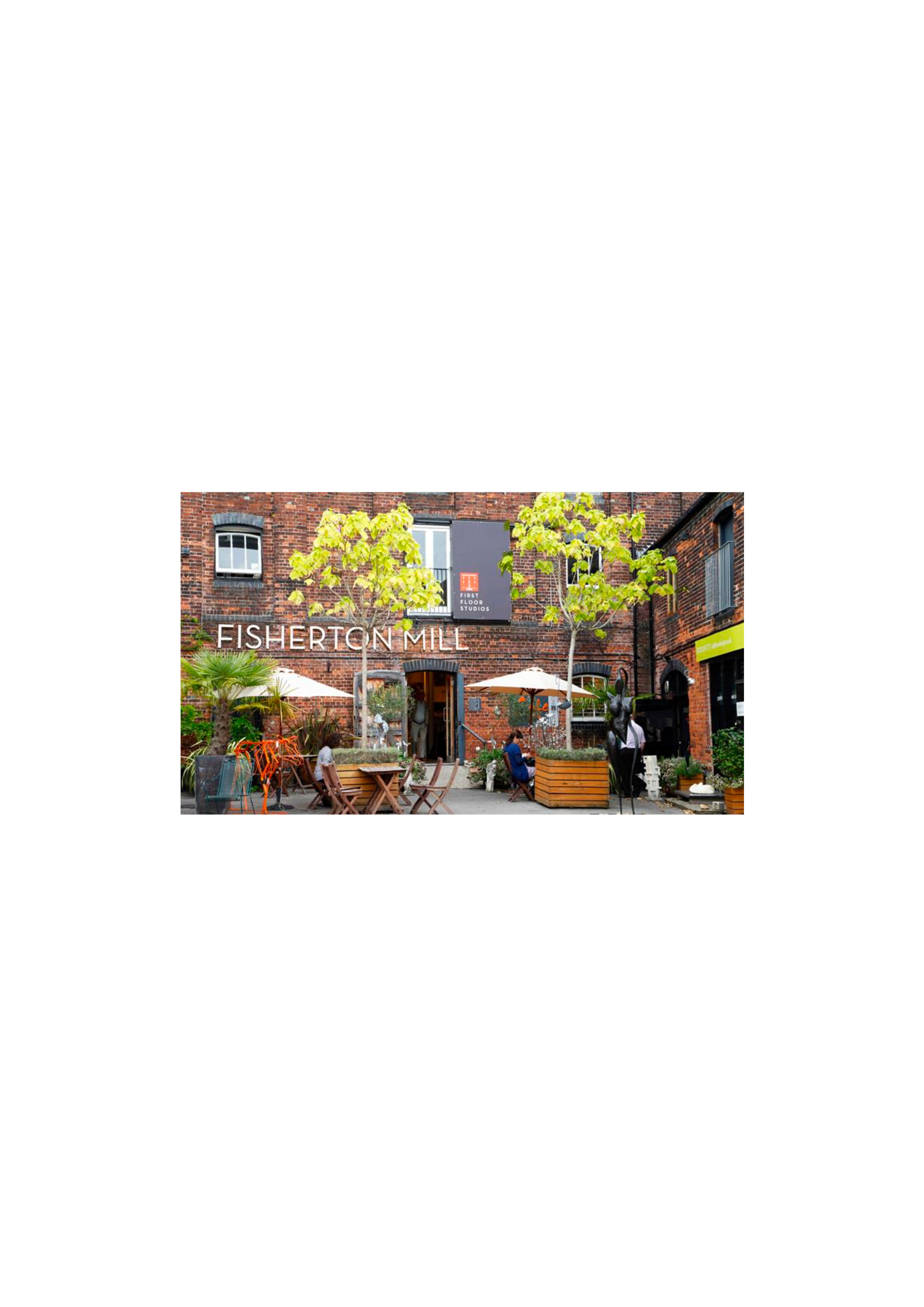 Outdoor patio area with wooden tables and chairs, green plants, trees, and a brick building with sign reading "FISHERTON MILL".