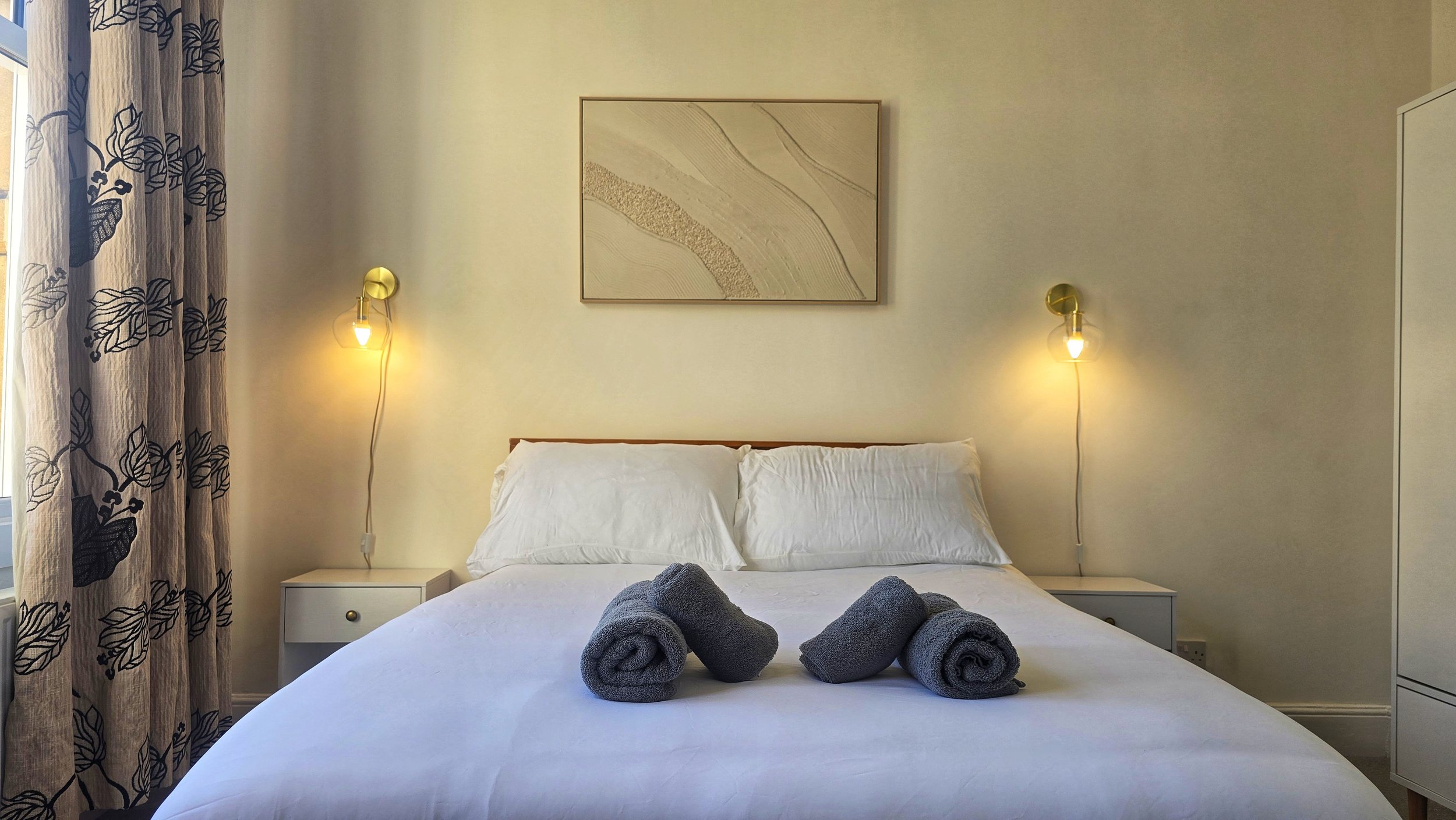 A neatly made bed with white bedding and two pillows, with three rolled gray towels on top. Two wall-mounted lamps are on either side of a framed abstract artwork on a beige wall. A white bedside table with a drawer is on each side of the bed. A window with curtains featuring black leaf patterns is on the left side of the image.