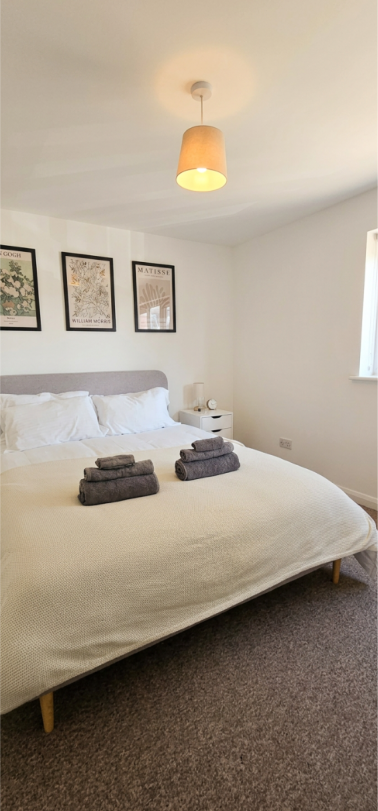 A neatly made bed with a beige bedspread and three rolled gray towels on top. The bed has a light gray headboard, and there are three white pillows. A white nightstand with a small lamp, clock, and other items is next to the bed. The wall above the bed has a series of framed art posters. A ceiling light with a beige lampshade hangs from the ceiling, illuminating the room.