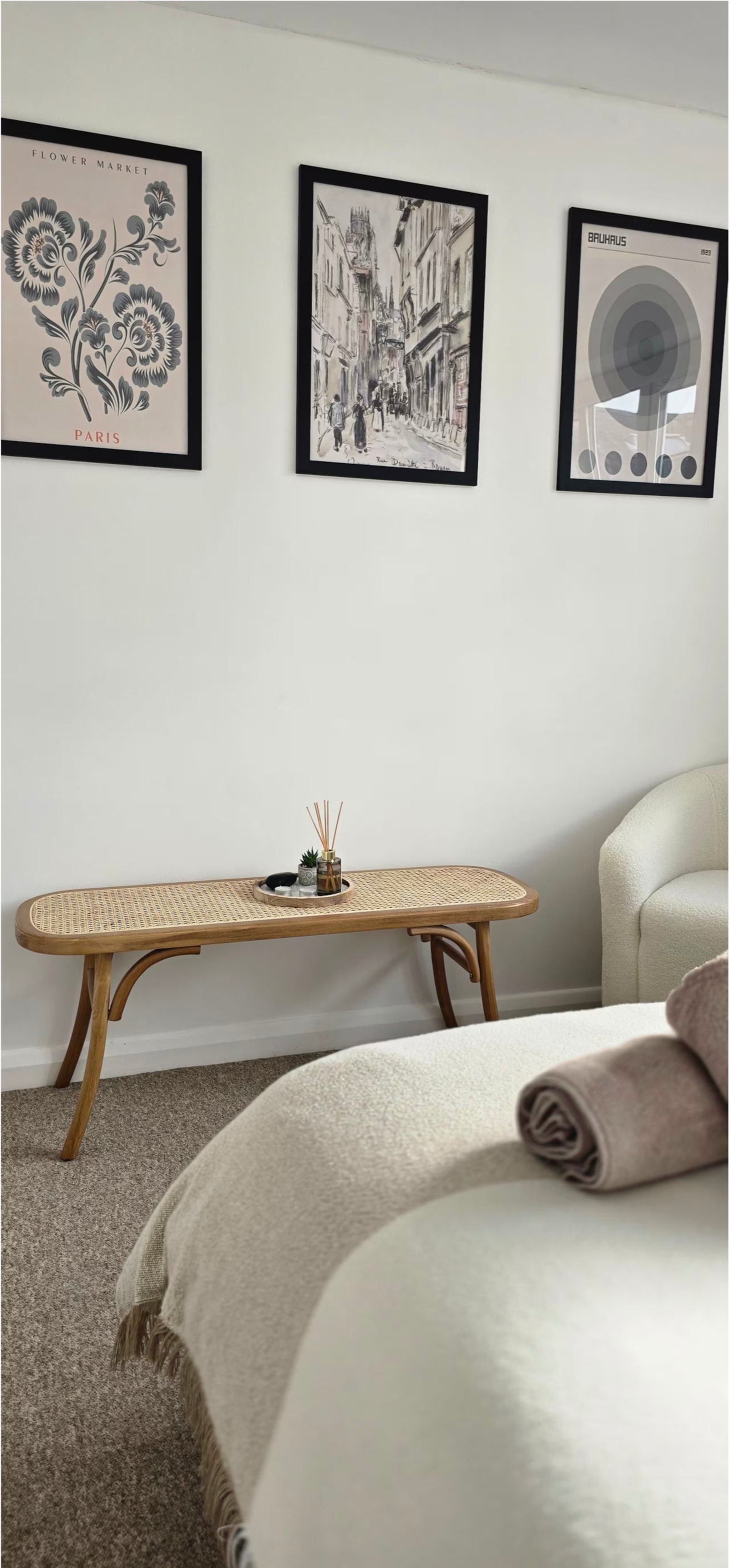 A cozy corner of a living room featuring a beige sofa with rolled towels, a wooden coffee table with decorative items, and three framed black-and-white art prints on the white wall.