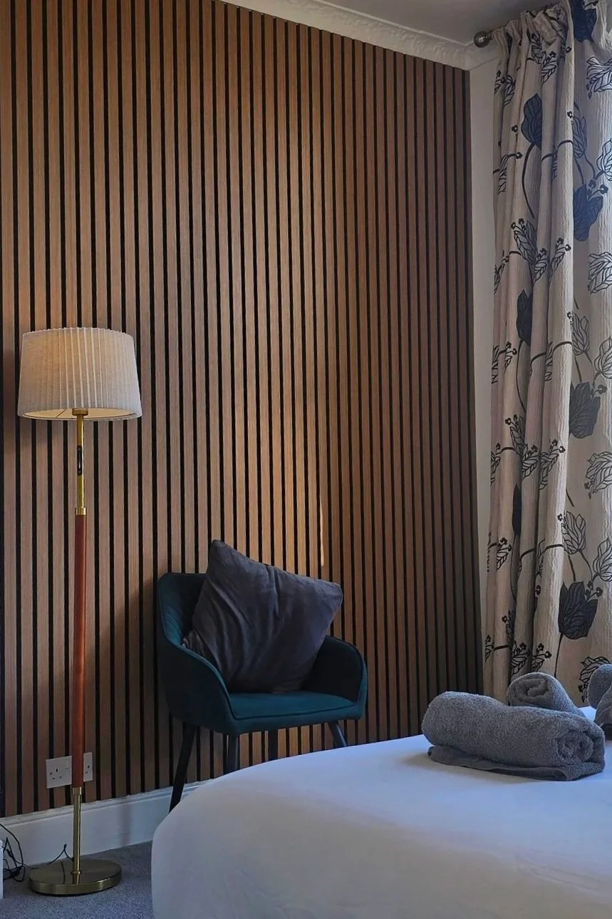 A bedroom corner featuring a wooden slat wall, a teal armchair with a dark cushion, a white bed with folded gray towels, a standing lamp with a beige shade, and a window with patterned curtains.
