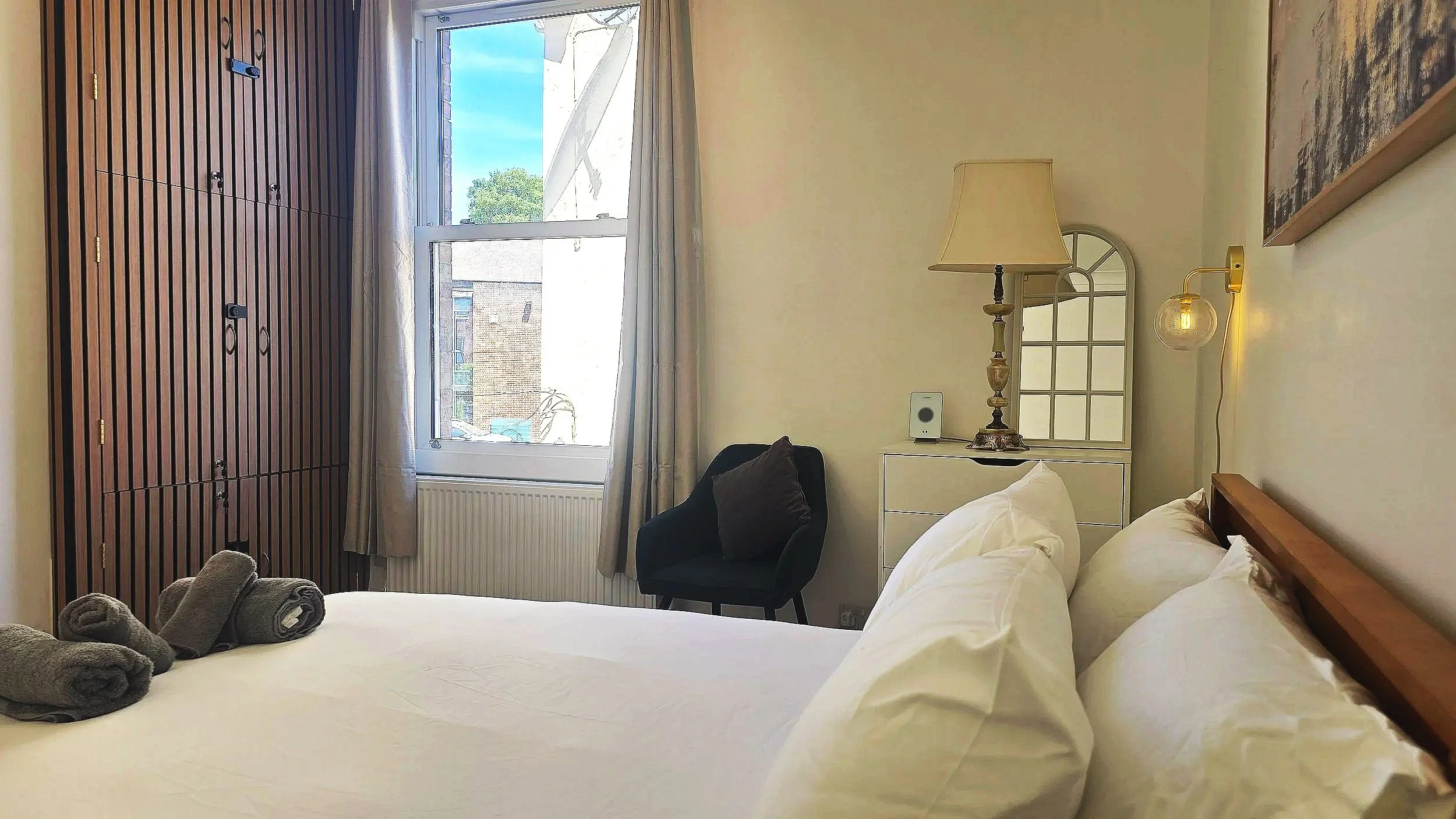 A cozy bedroom with a large bright window, a bed with white bedding, rolled towels on the bed, a black armchair with a pillow, a white nightstand with a table lamp, and wall art. The wooden wardrobe has a slatted design, and there's a small mirror on the wall.