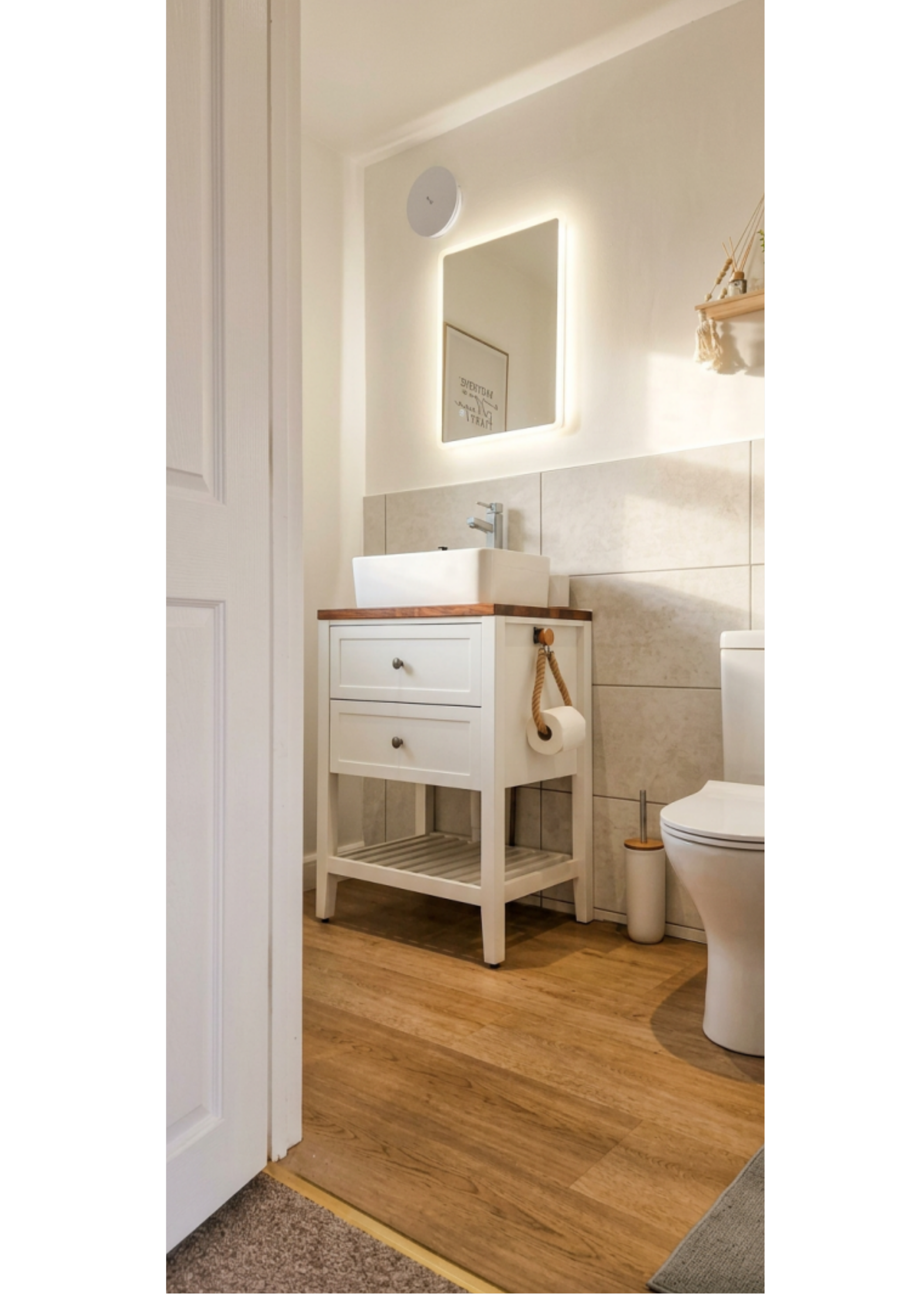 A bathroom with white walls and a wooden floor. There is a small white cabinet with a sink on top, a framed quote on the wall with a mirror, a shelf with decorative items, a toilet with a toilet paper holder, and a wooden toilet brush. The door frame is visible on the left side, and a section of beige carpet is visible in the foreground.