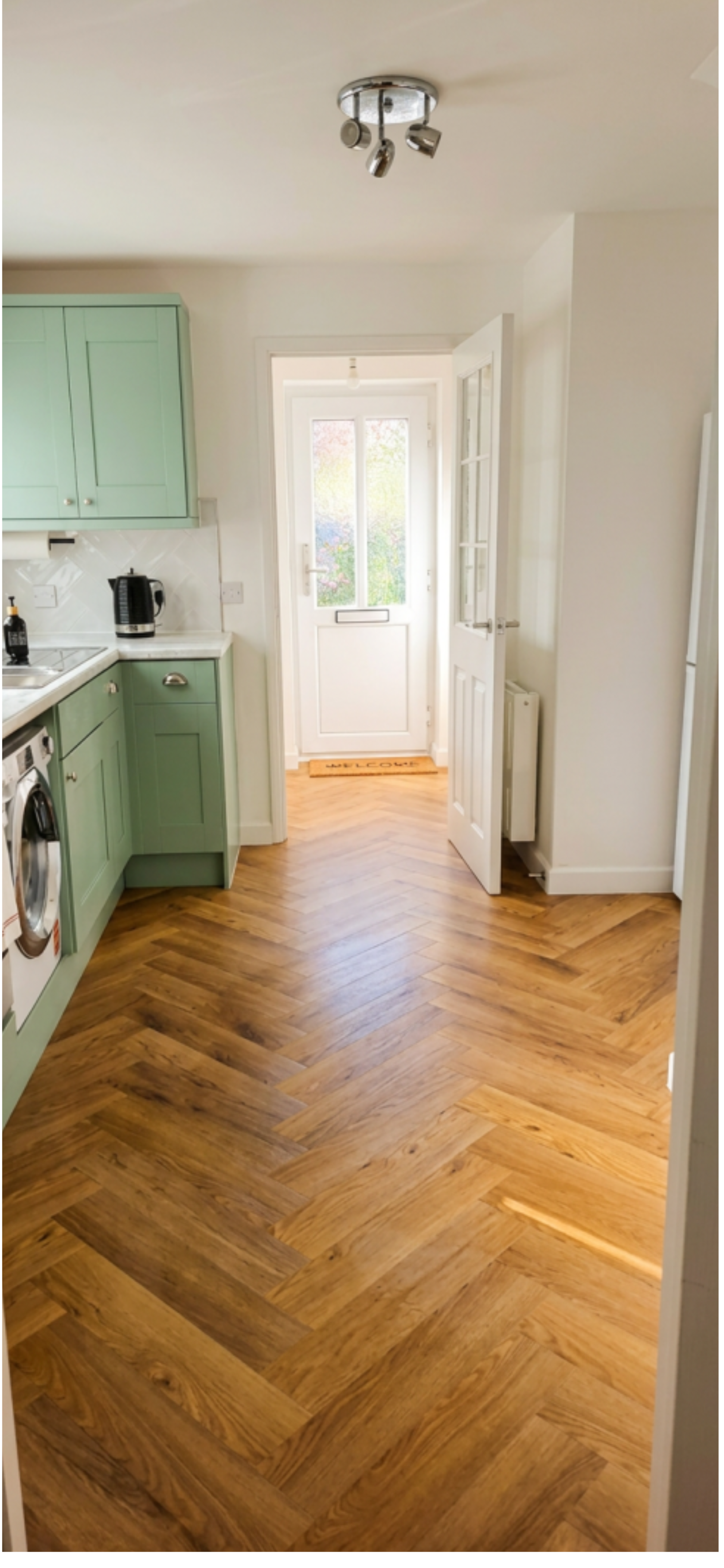 A kitchen with green cabinets, wooden herringbone-patterned floor, and a white door leading outside with a welcome mat.