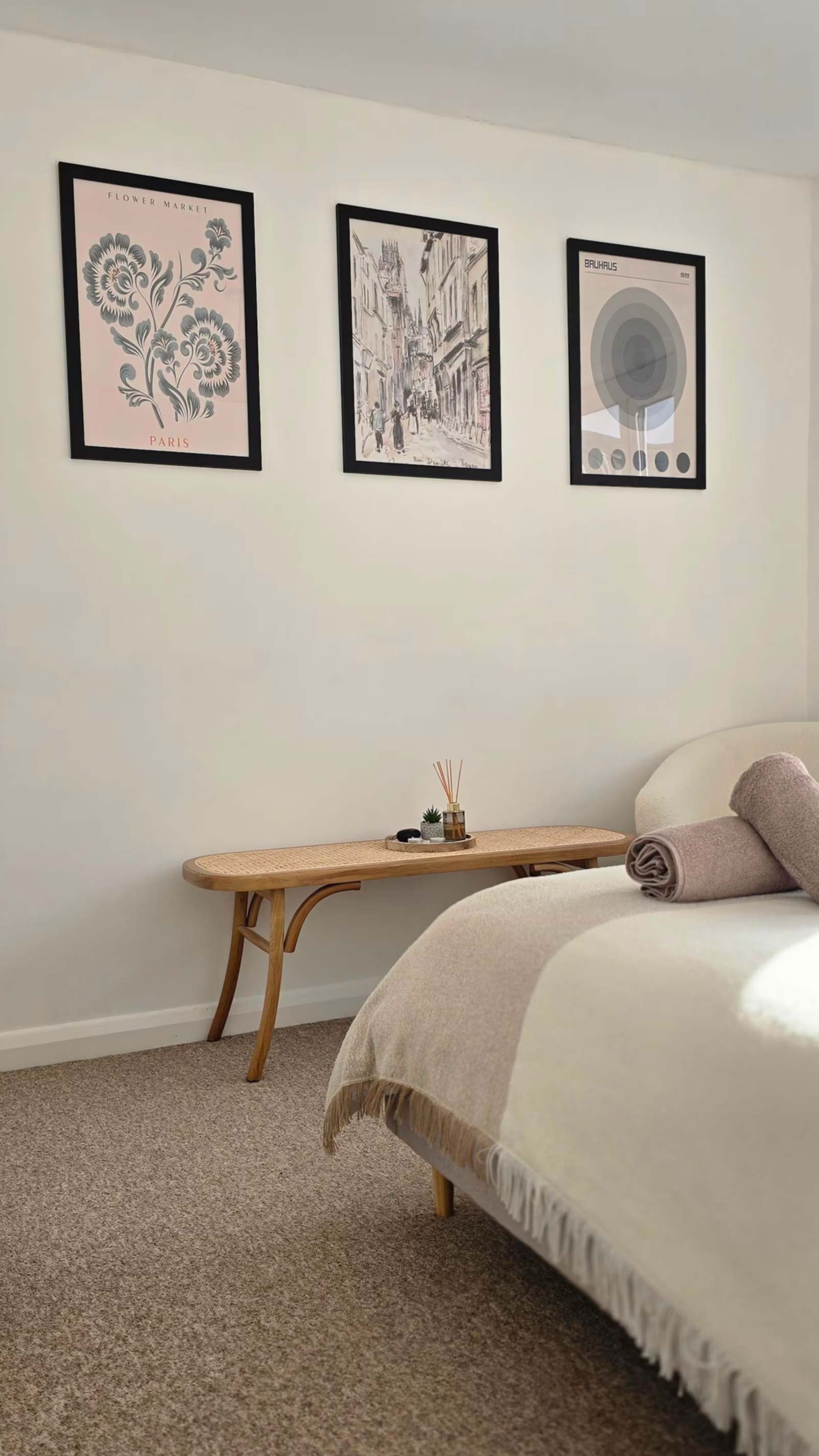 A cozy bedroom corner with a bed covered in a beige blanket and rolled towel. A wooden side table with small decorative items is against a white wall, which displays three framed art prints.