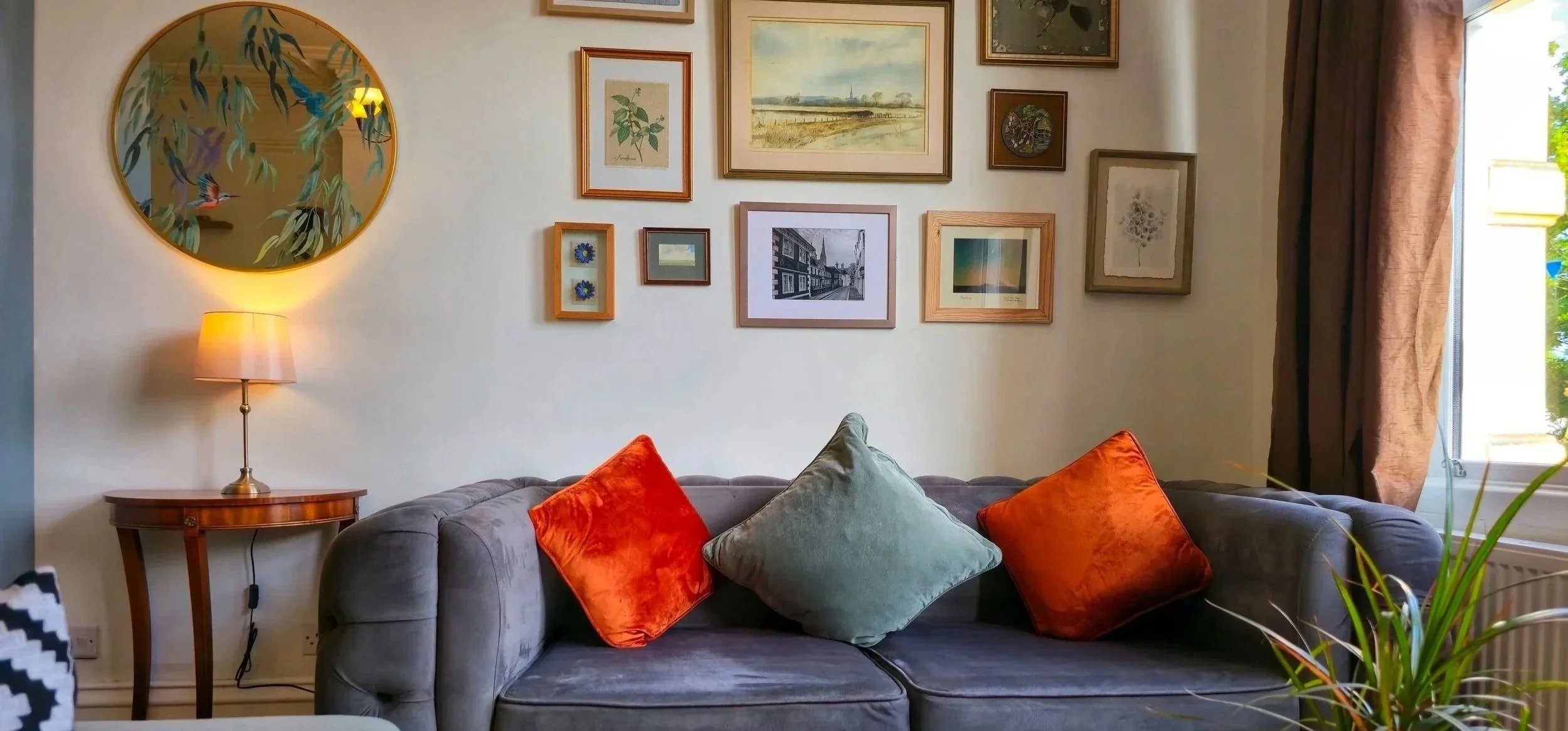 A living room with a gray sofa decorated with green and orange throw pillows. A wooden side table with a lamp stands beside the sofa. A gallery wall with various framed artworks and photographs is behind the sofa. A window with brown curtains is on the right side of the image, allowing natural light to enter.