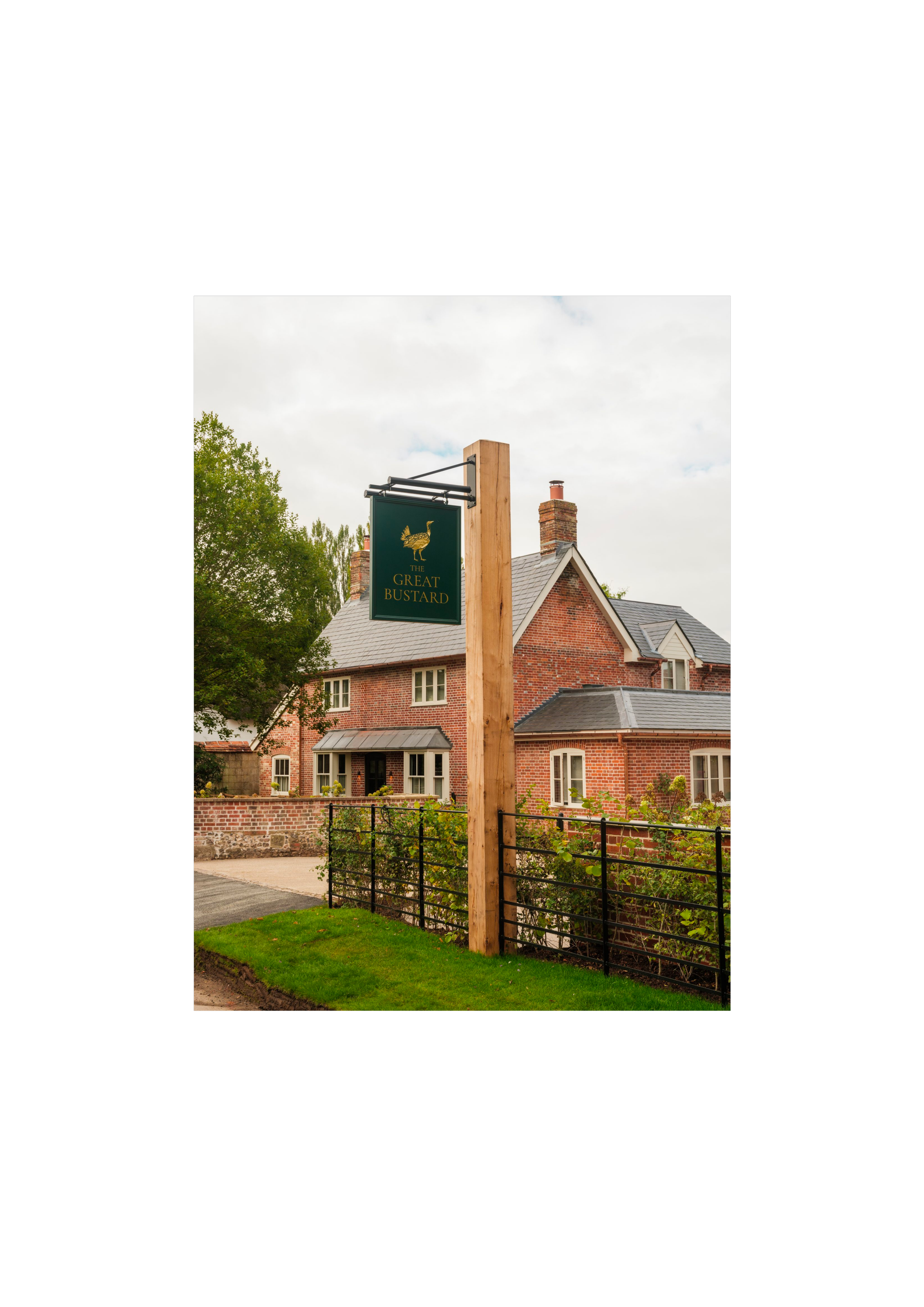 A sign hanging from a wooden pole reads 'The Great Bustard' with a bird illustration on a green background, in front of a red brick house with chimneys and a garden.