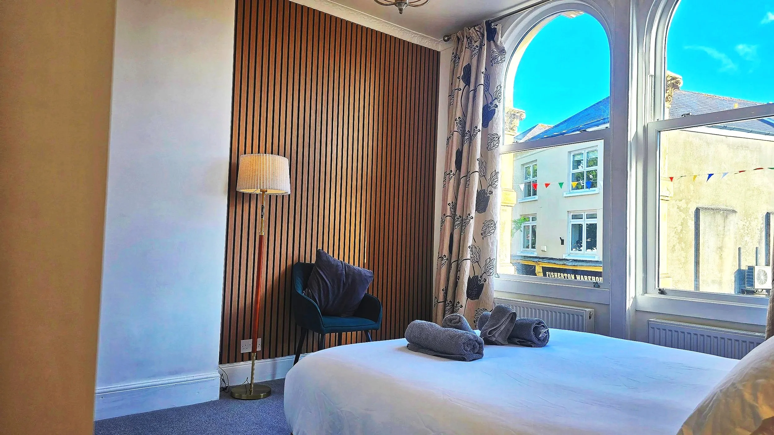 A bedroom with a large window showing a building and blue sky outside. The room has a wooden slat accent wall, a standing lamp with a cream shade, and a black chair with a pillow. On the bed, there are rolled towels.