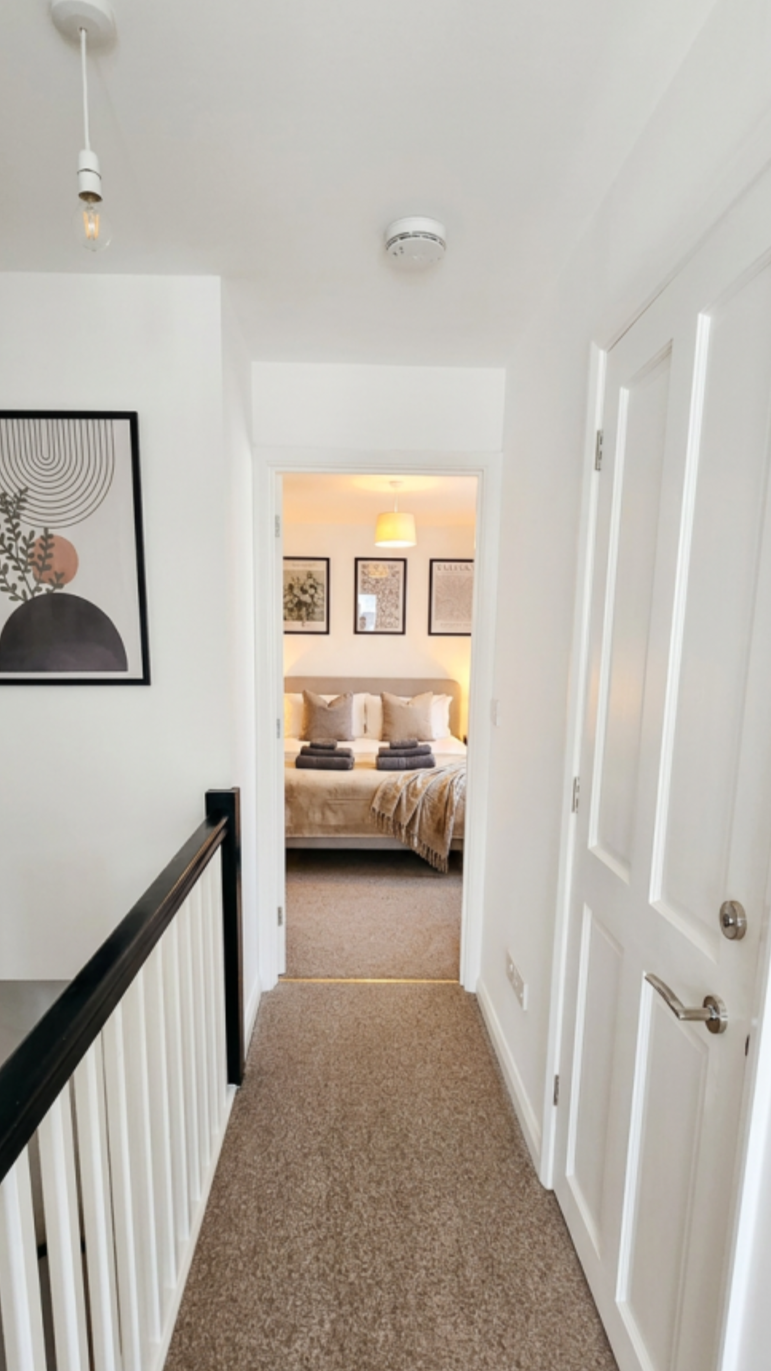 View of a hallway leading to a bedroom with a bed and framed pictures on the wall, hardwood railing on the left, white walls, beige carpet, ceiling smoke detector, and a door on the right.