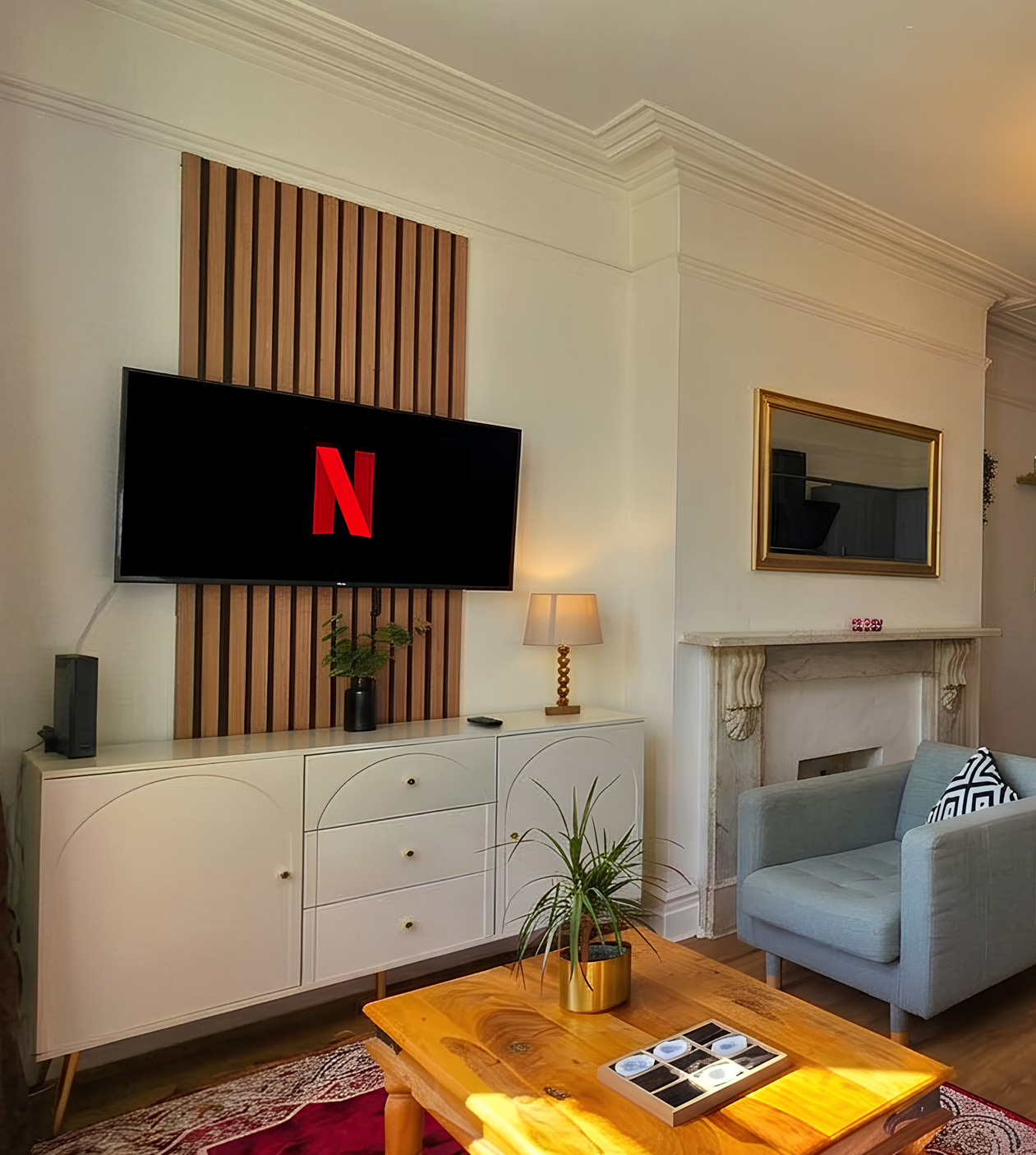 Living room with a mounted flat-screen TV displaying the Netflix logo, a sideboard, a lamp, a mirror above a fireplace, a plant on a wooden table, and an armchair with a cushion.