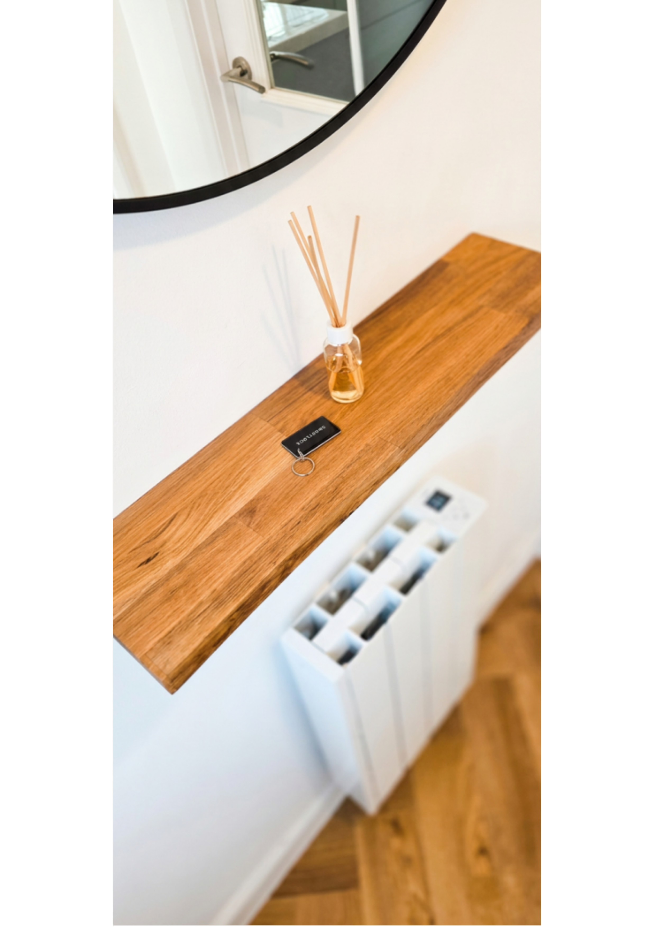 A wooden shelf on a white wall with a glass reed diffuser and a small black keychain. A mirror reflecting a door handle is visible above the shelf.