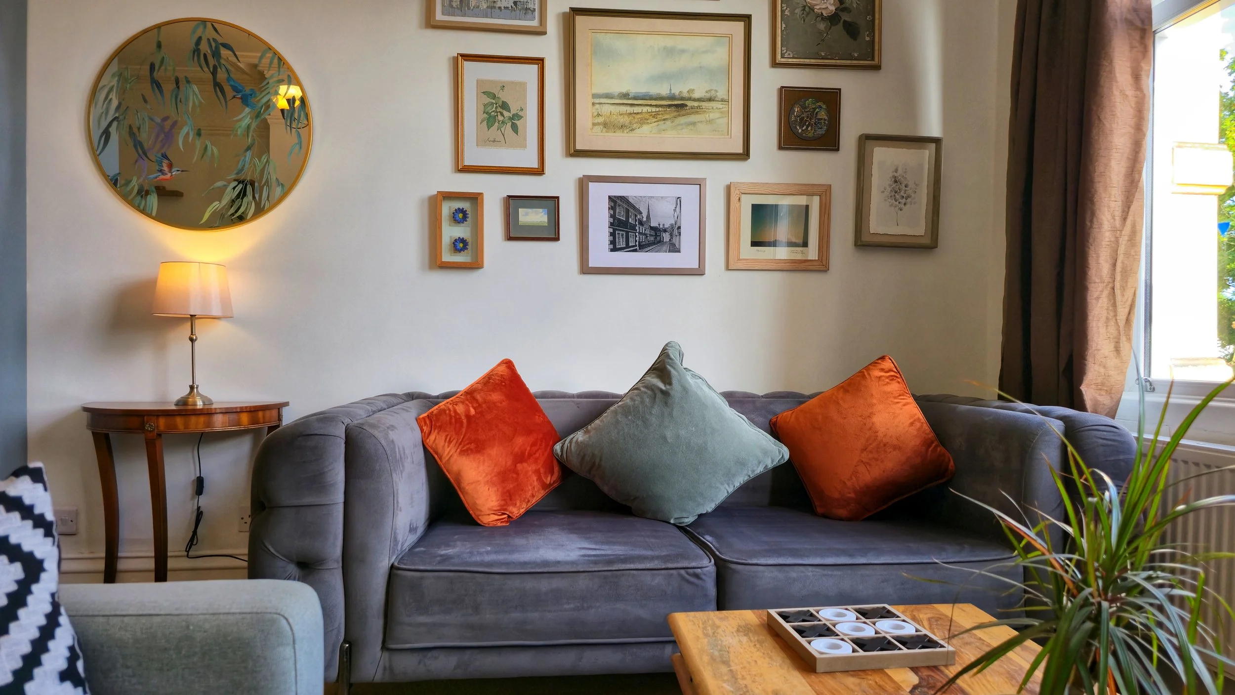 A cozy living room with a gray sofa decorated with four colorful pillows, a wooden side table with a lamp, and a gallery wall of framed artwork and photographs above the sofa. Part of a window with curtains is visible on the right side.