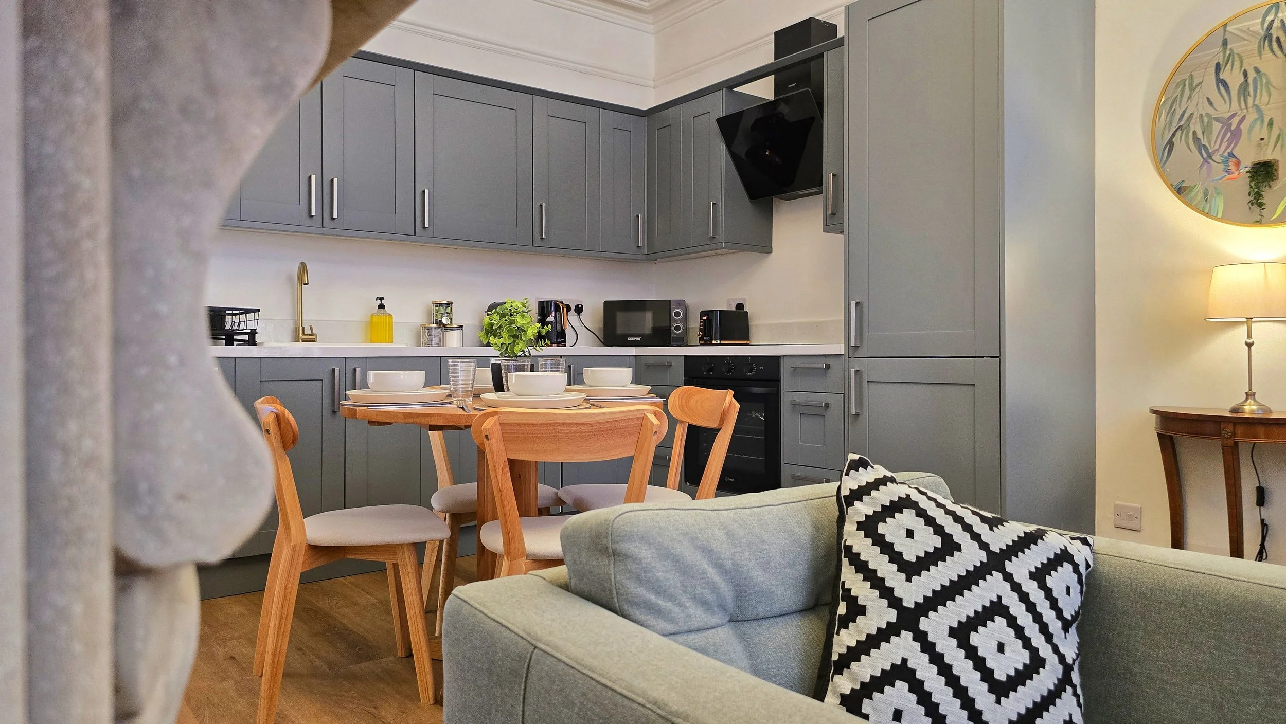A cozy kitchen and dining area with a round wooden table set with bowls, glasses, and a plant, surrounded by four wooden chairs, adjacent to a living space with a green sofa and a black and white patterned pillow. The kitchen features gray cabinets, black appliances, a microwave, and a small window with a yellow lampshade nearby.