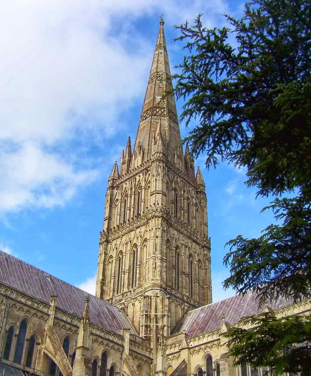 alisbury Cathedral Gothic spire - a short walk from Tirgo Collection holiday lets in the city centre. A tall, historic cathedral with a Gothic-style spire against a blue sky with clouds, partially framed by trees.