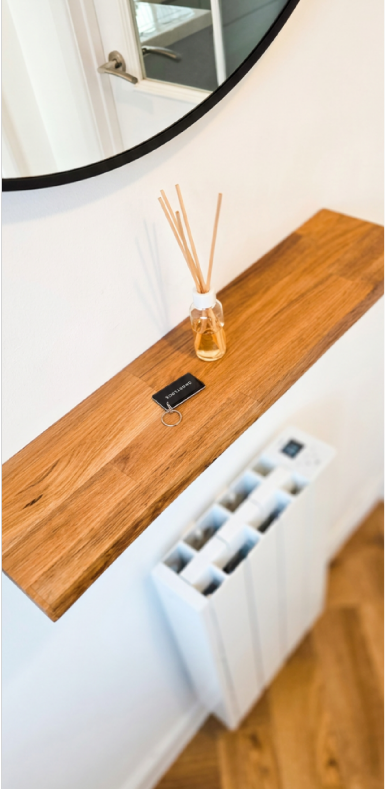 Wooden ledge with a diffuser bottle with reed sticks, a black keychain, and a white radiator below. A mirror and door with a handle reflect a corner of the room.