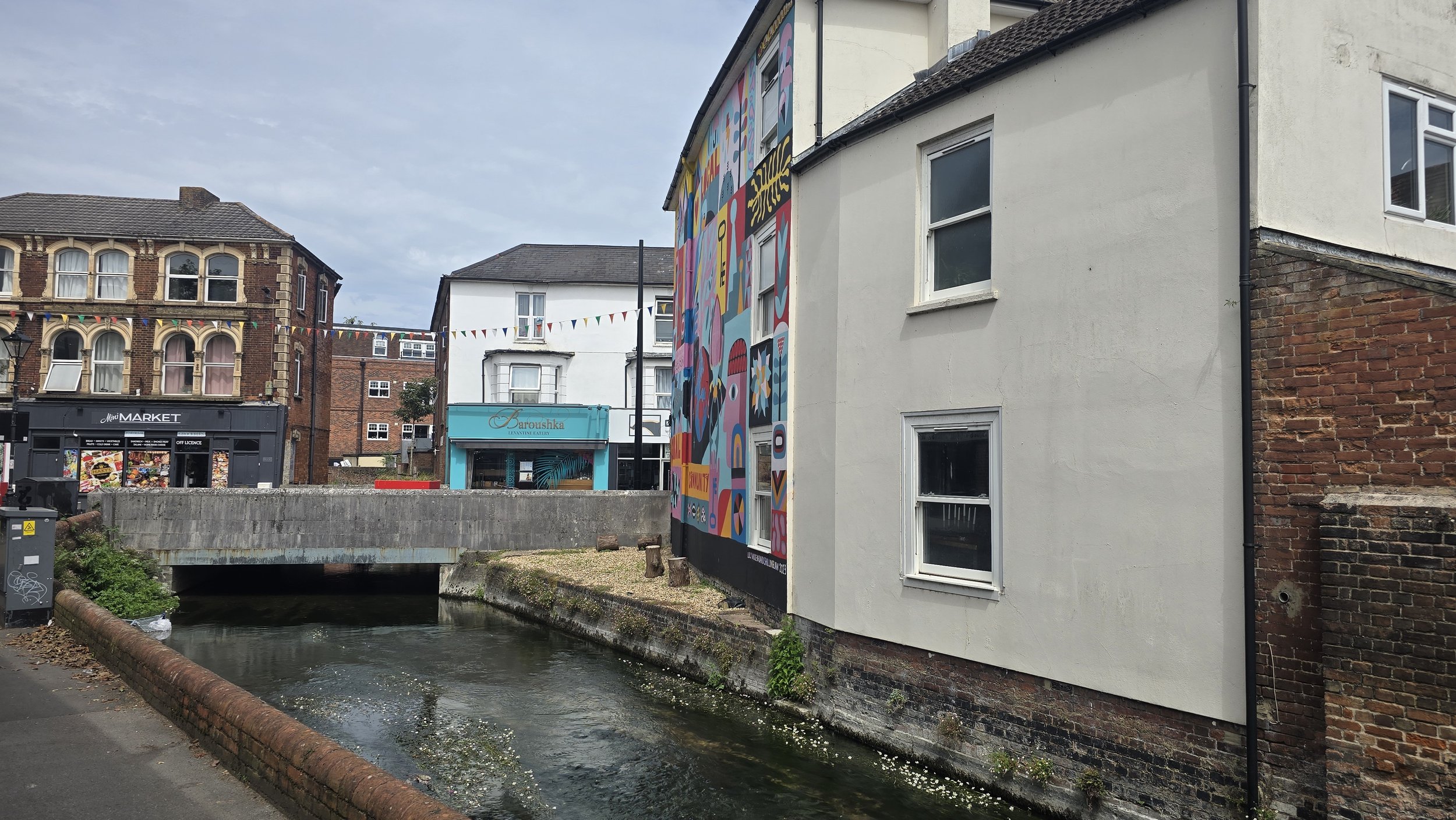 A canal with water flowing under a bridge, lined with buildings including a brick building and a white building with a large colorful mural on its side. There is a shop named 'Mini Market' and another with a blue sign, 'Baroushka Lounge & Eatery.' Colorful bunting is strung across the street.