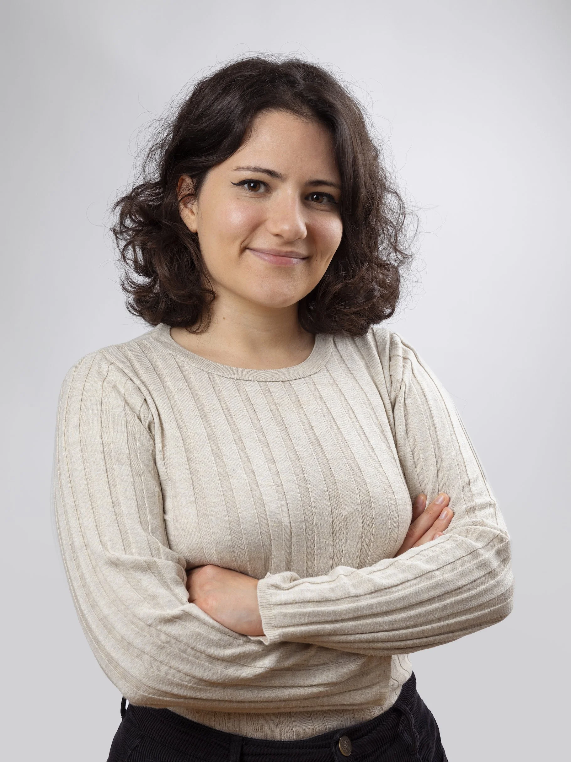A young woman with dark brown, shoulder-length curly hair and light skin, standing with her arms crossed and smiling slightly, against a plain gray background, wearing a beige ribbed long-sleeve sweater and black pants.