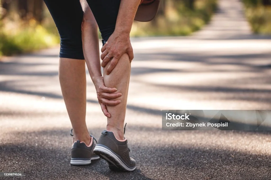 Person holding their sore calf muscle while standing on a paved road outdoors.