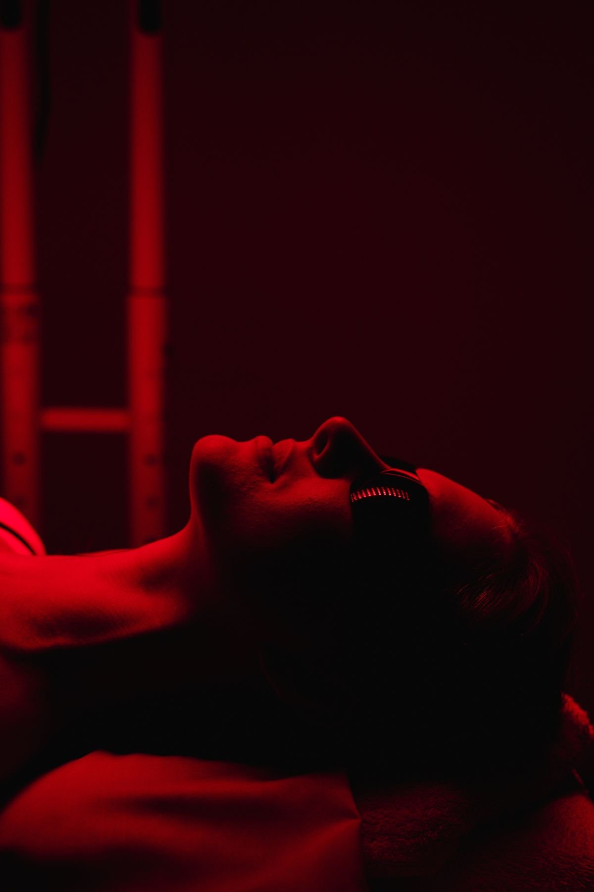 A woman lying face down on a bed, illuminated by red lighting from a heat or infrared lamp overhead, with her head turned to the side, wearing a headband.