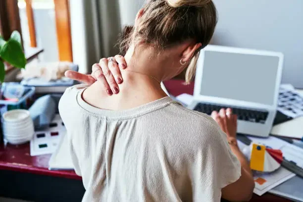 A woman with her back to the camera experiencing neck pain while sitting at a desk with a laptop and various office supplies.