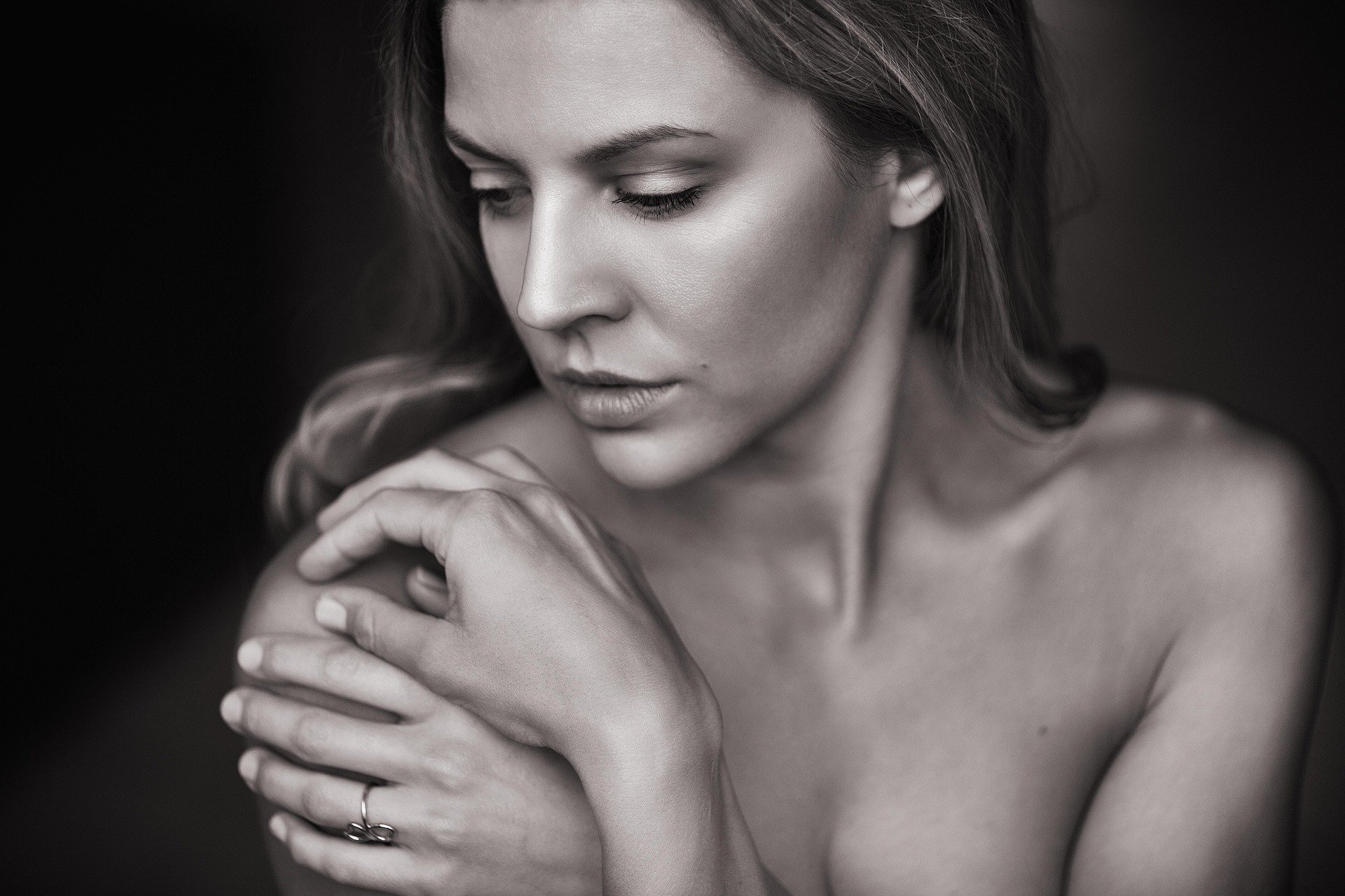 Black and white portrait of a woman with closed eyes, gently touching her shoulder with her hands, minimal makeup, soft lighting.