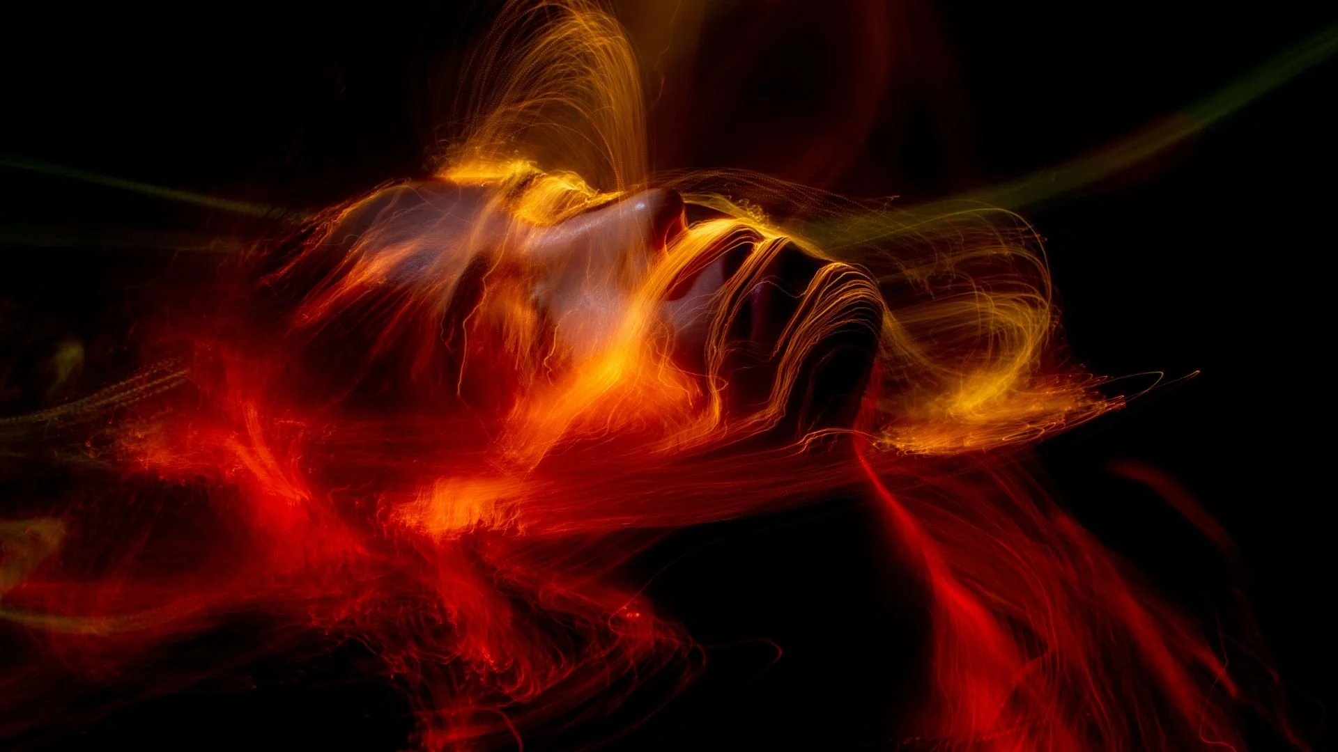 Abstract long-exposure photograph of a person's face and hands created with fiery light trails in reds, oranges, and yellows against a dark background.