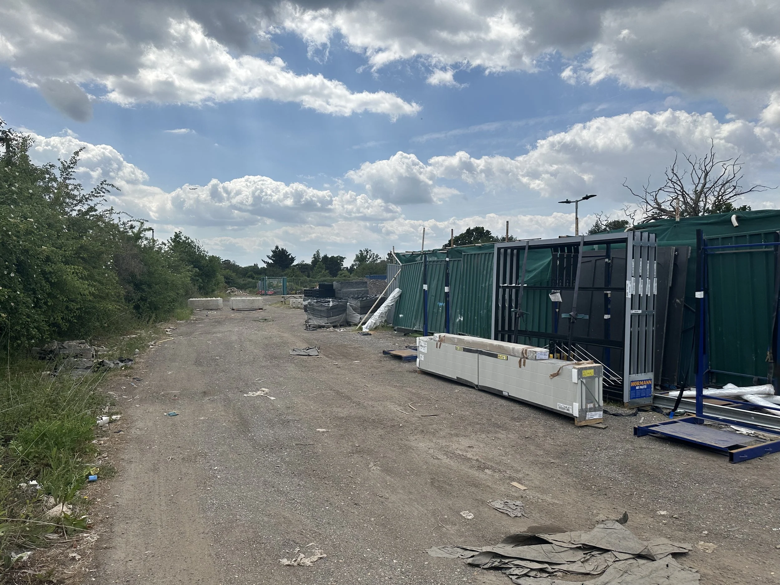 A partially clear sky with scattered clouds over an outdoor area with construction materials and debris, including metal fencing, pallets, and miscellaneous objects, surrounded by green bushes and trees.