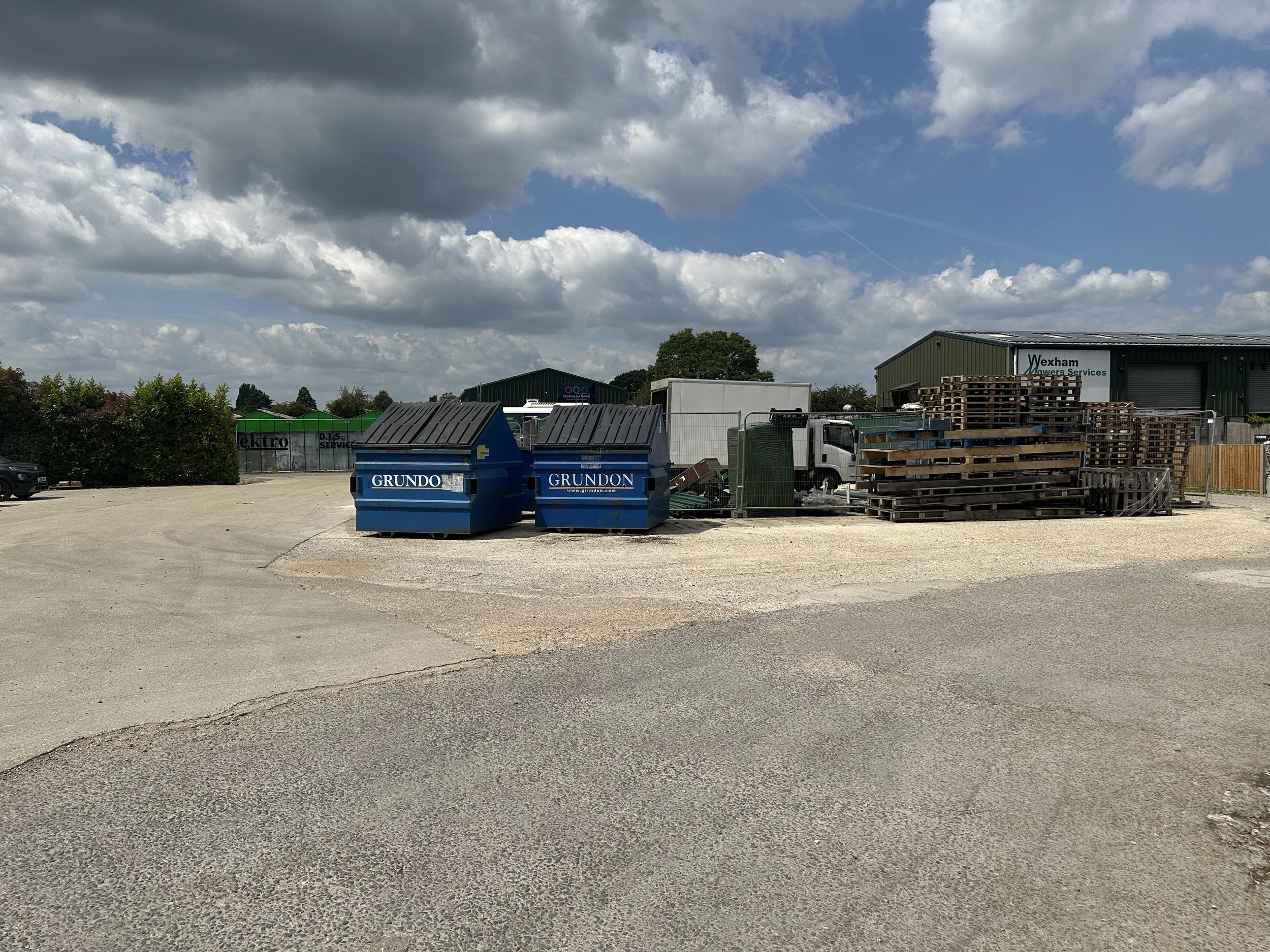 A parking lot with gravel surface, blue dumpsters labeled 'Grundon', and a green industrial building with a sign 'Wexham Movers Services'. There are stacked wooden pallets and a few vehicles, with cloudy blue sky overhead.