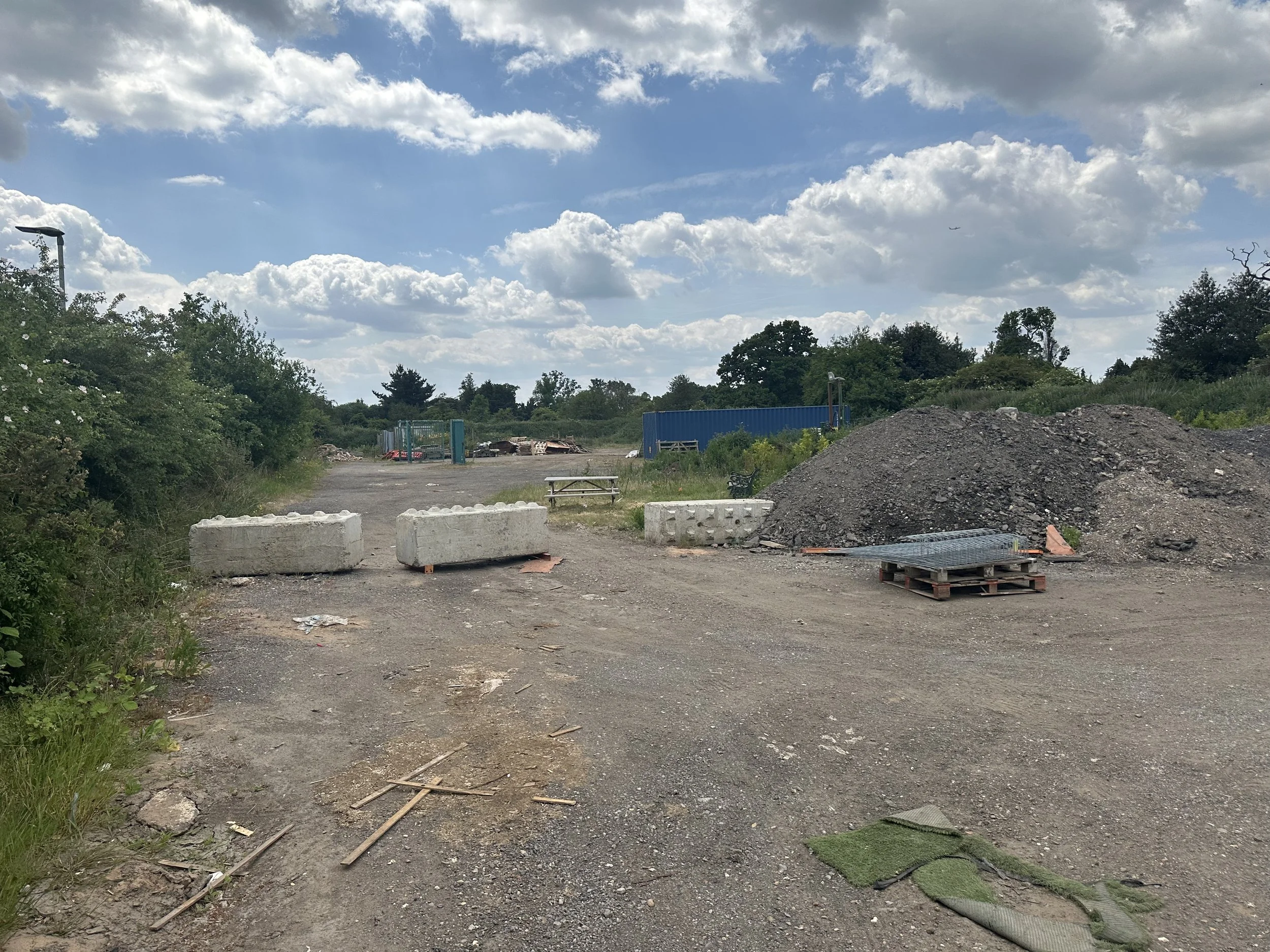 An outdoor scene with a dirt and gravel area, some trees and bushes, a large pile of dirt, and construction materials. The sky is partly cloudy with some clouds and a plane flying in the distance.