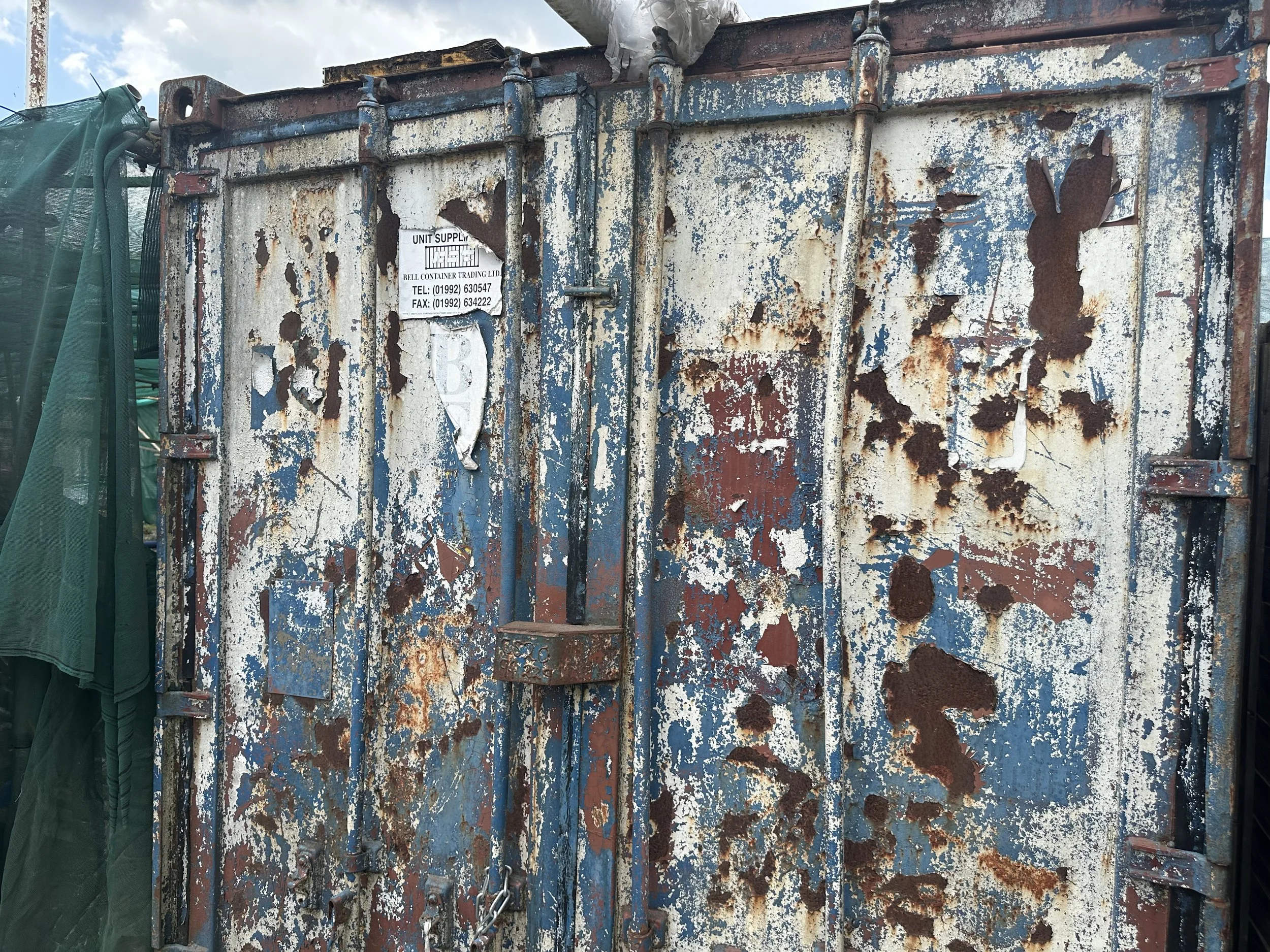 An old, rusty metal container with peeling blue and white paint, chained shut with a padlock. There is a partially torn white sign with black text on the container.