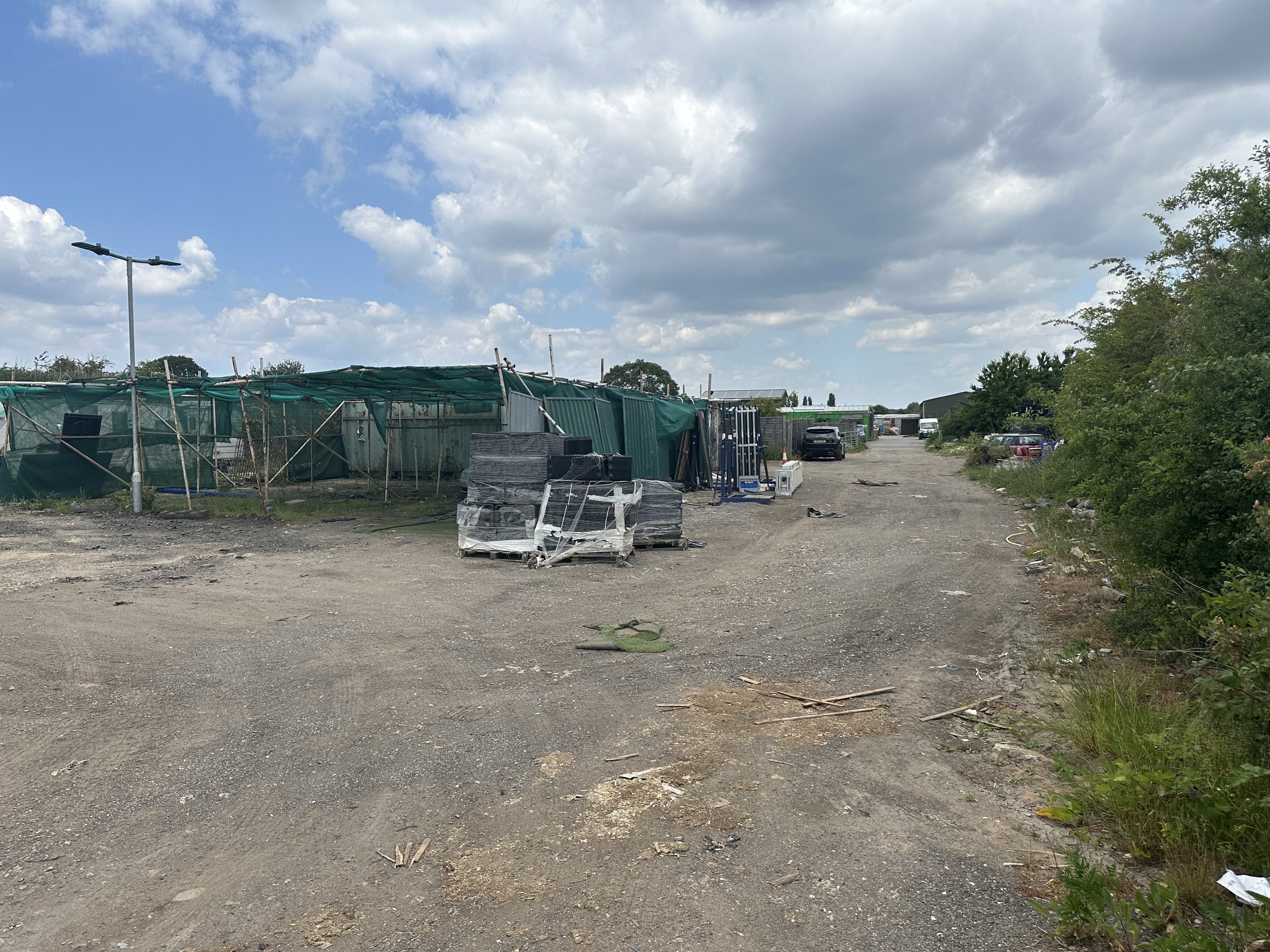 A gravel lot with scattered debris, patches of grass, construction materials, and a green fabric barrier. There are several parked cars, trees, and a cloudy sky overhead.