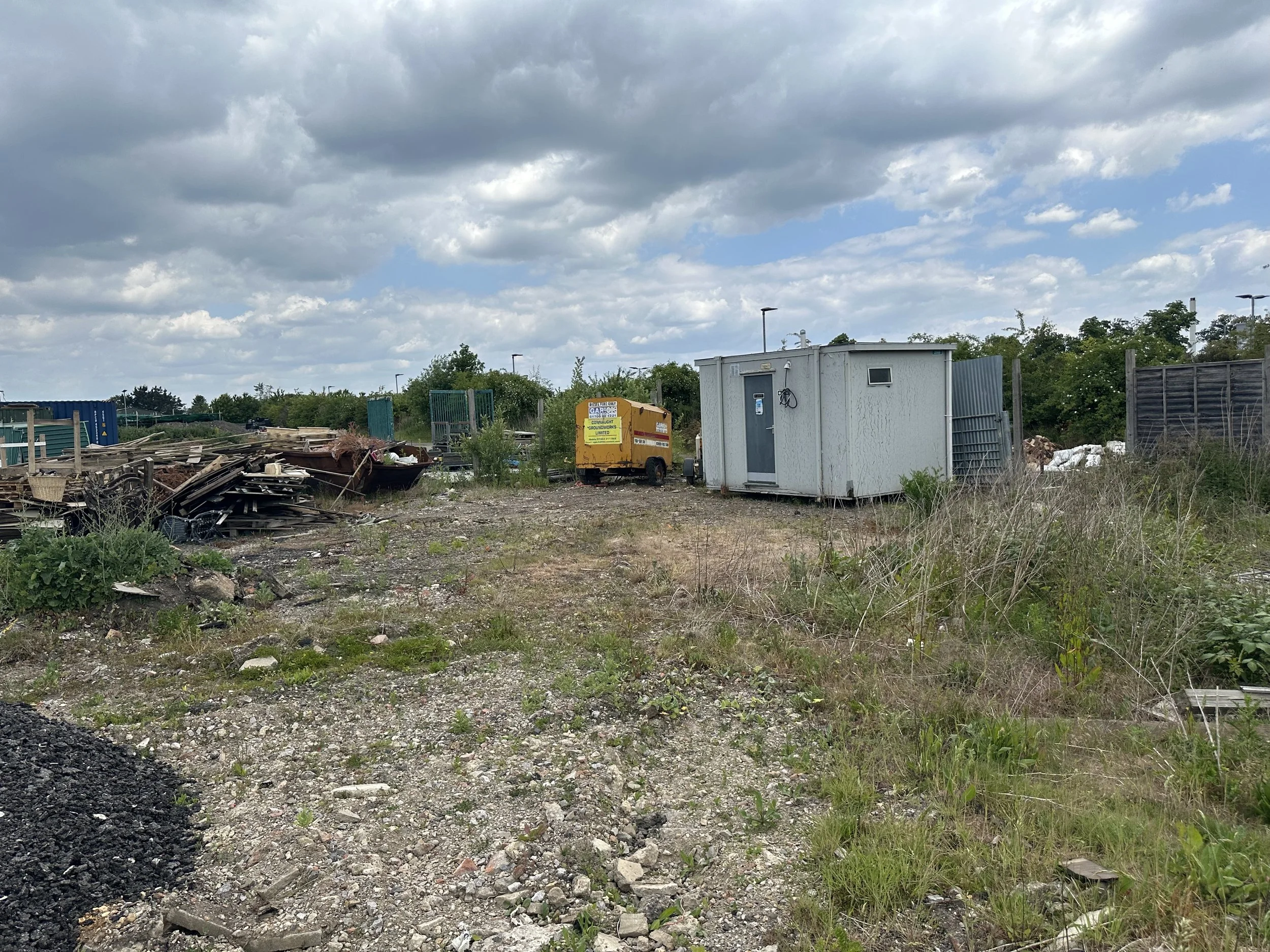 An outdoor scene with a partly cloudy sky, some greenery, and a gravel area containing debris, a portable building, and a yellow generator.
